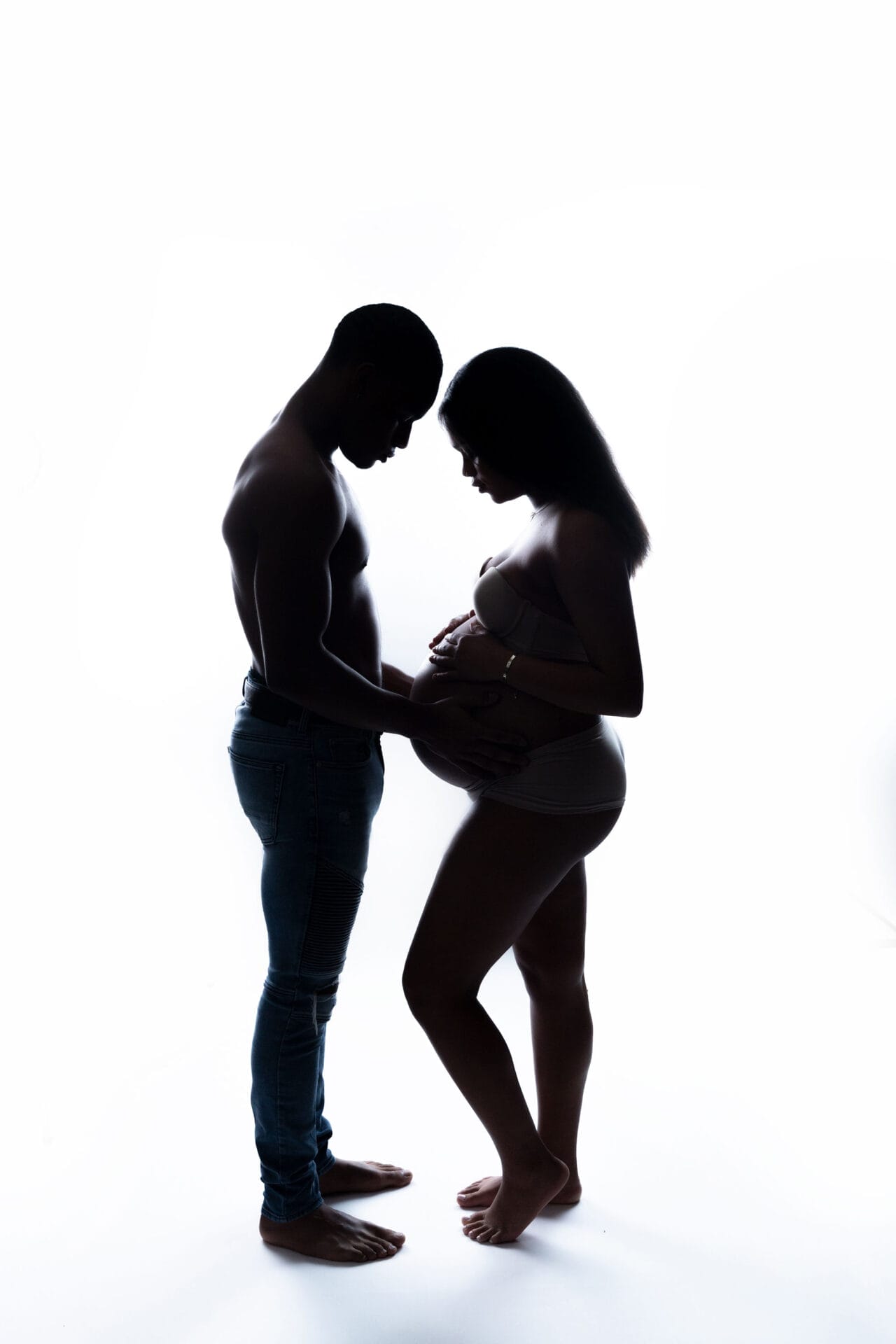 A silhouette of an Expecting couple in the studio for their maternity face-to-face session, standing, embracing her pregnant belly.