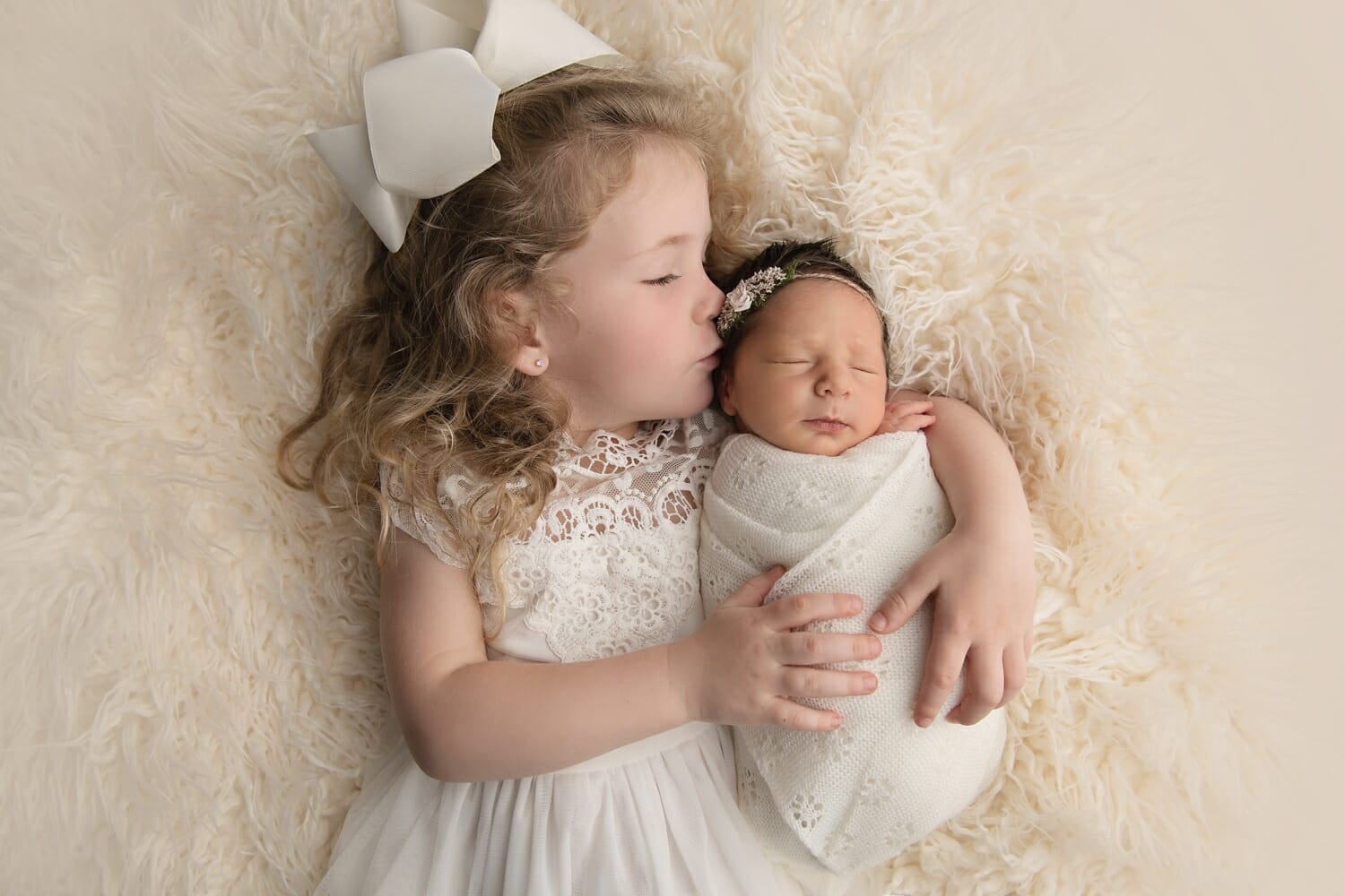 A sibling lays beside a newborn baby on a white rug
