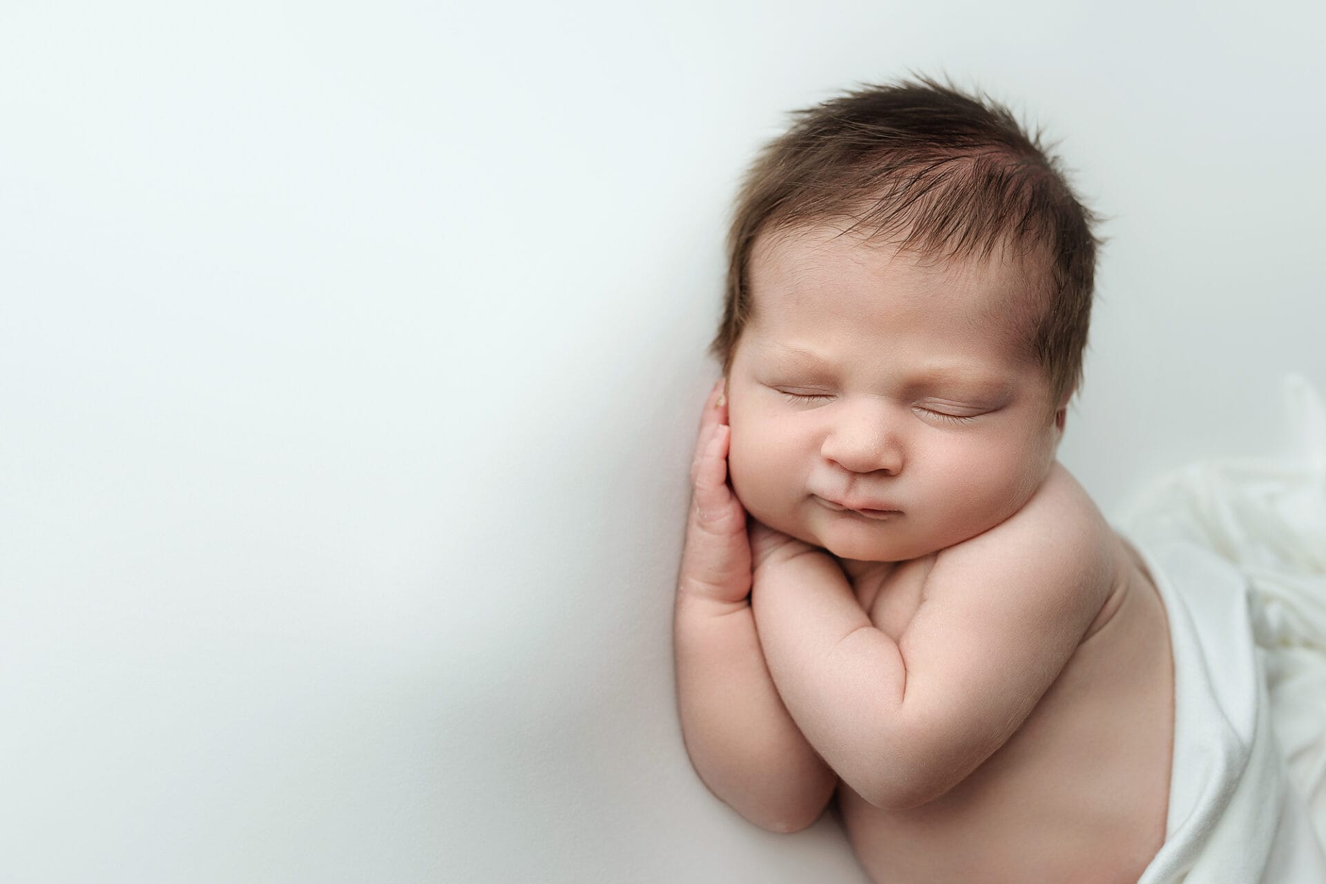 A newborn baby boy lying on a white backdrop with hands under his cheek.