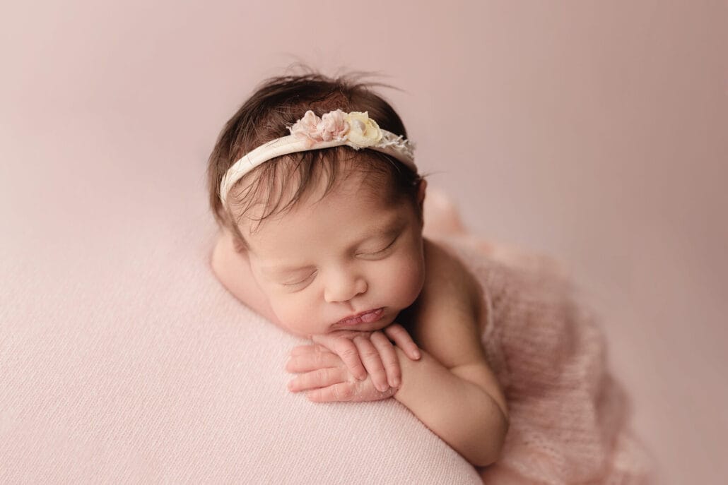 A newborn baby girl on a pink backdrop with a hand under the chin with a floral headband. 