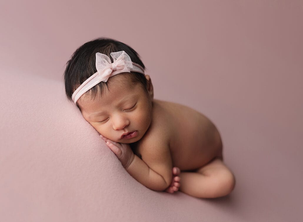 This is a photo of a baby girl on a pink backdrop in a taco pose with a petite headband and a pink bow with New Orleans newborn photographer. 