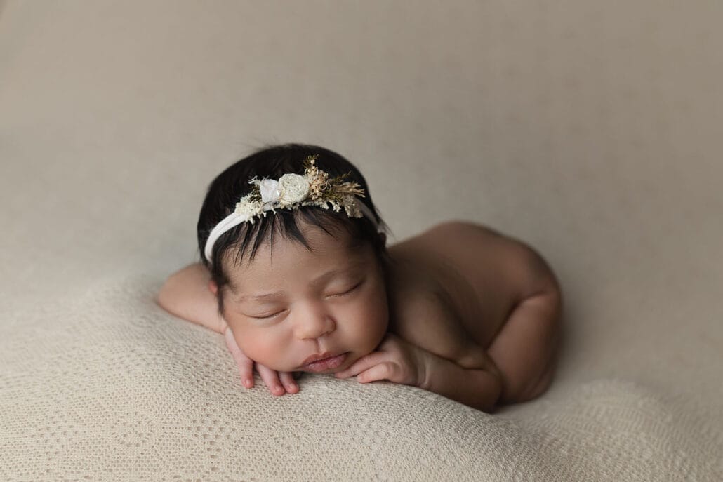 Newborn baby girl laying on tan backdrop on tummy with hand under chin wearing a floral head band with New Orleans childbirth class. 
