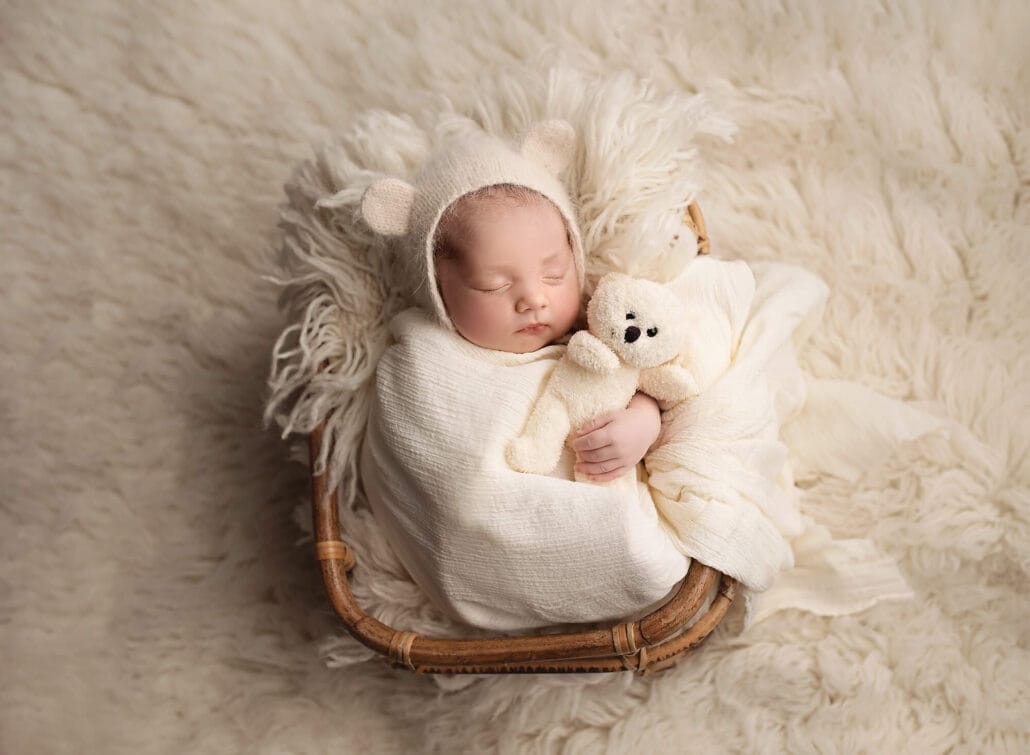 The newborn baby with bear bonnet is in the studio in a wicker basket and swaddled, cuddling a bear.