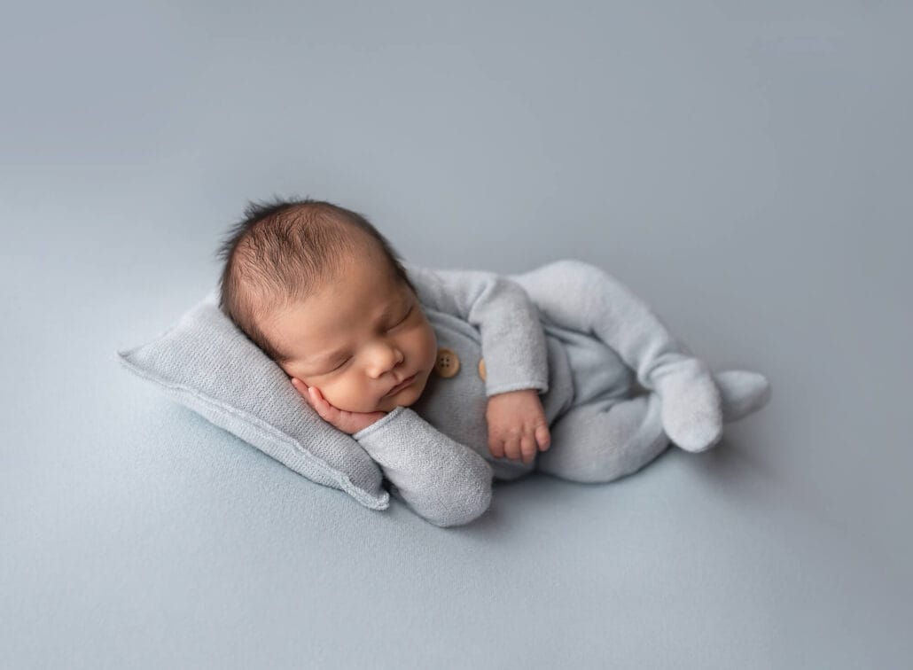 Baby boy in blue-footed outfit lying on blue backdrop with a pillow in studio.