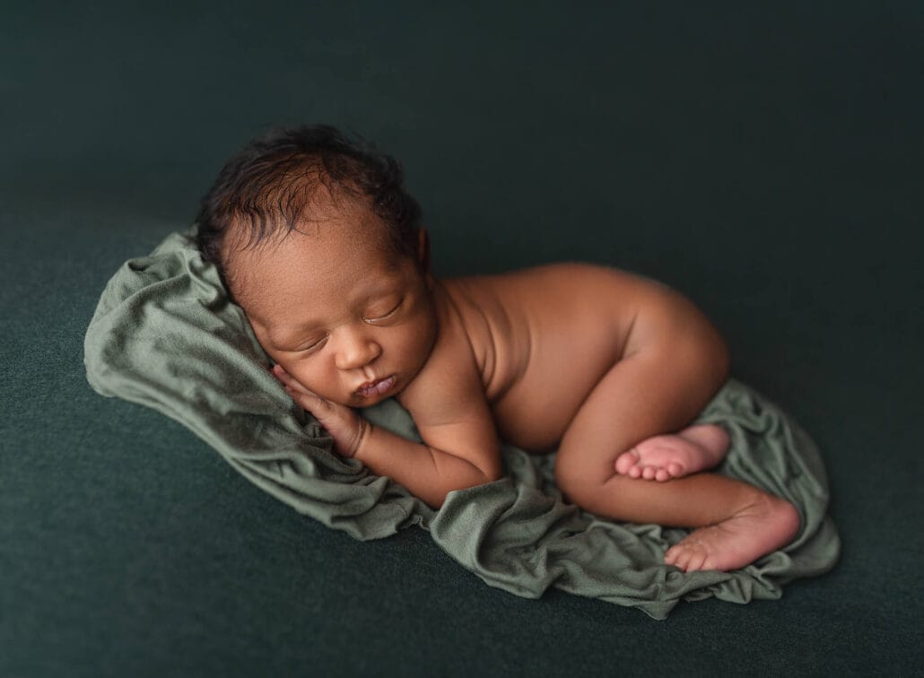 Newborn baby lying on green backdrop with feet curled and hand under chin.