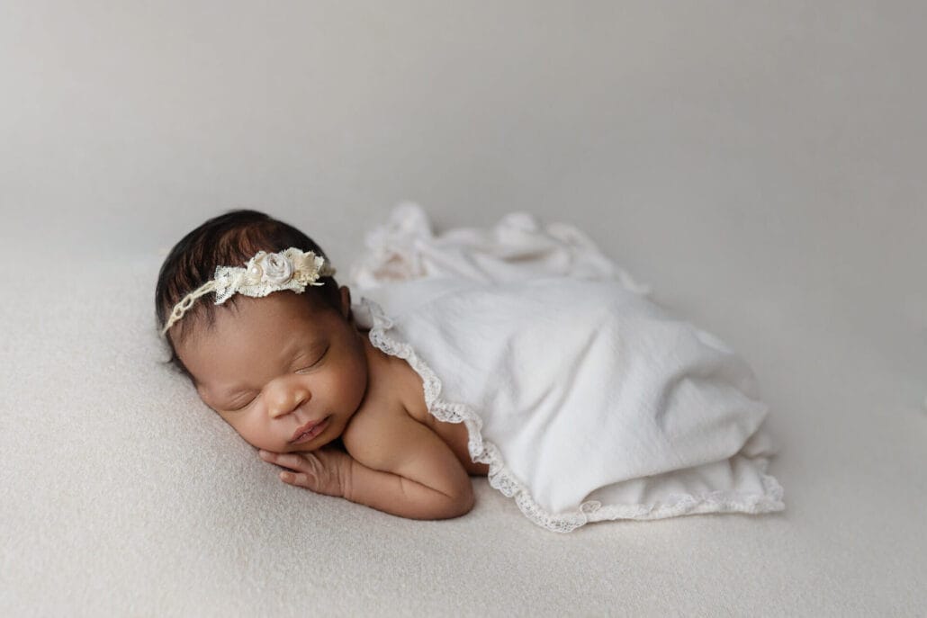 Newborn baby girl with ivory floral headband lying on cream backdrop with lace wrap in portrait studio.