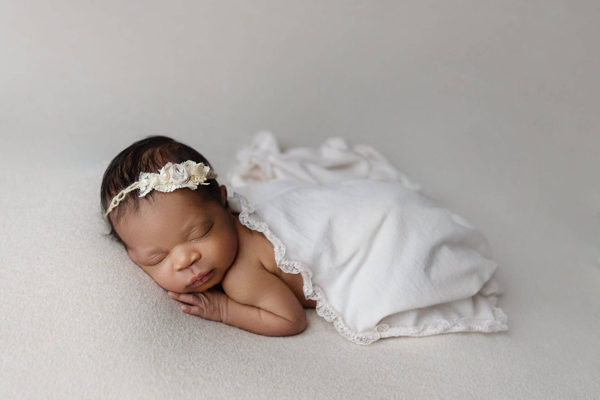 Newborn baby girl with ivory floral headband lying on cream backdrop with lace wrap.