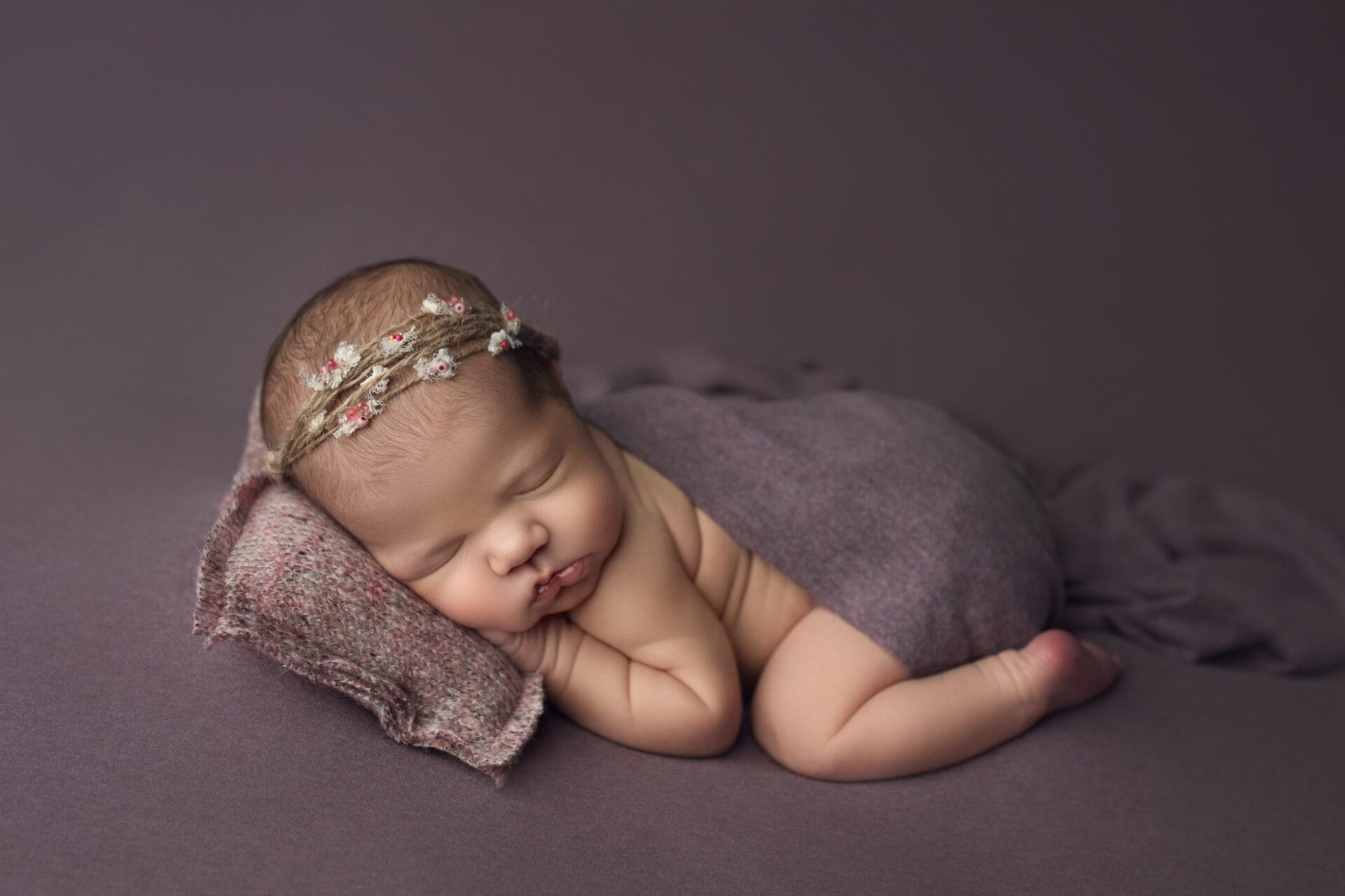 A newborn baby girl cuddled on the purple backdrop with a purple pillow.