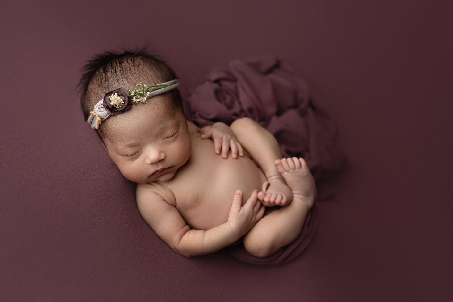 Newborn baby girl in womb poses with wine-colored headband and backdrop with best Baton Rouge baby boutique.