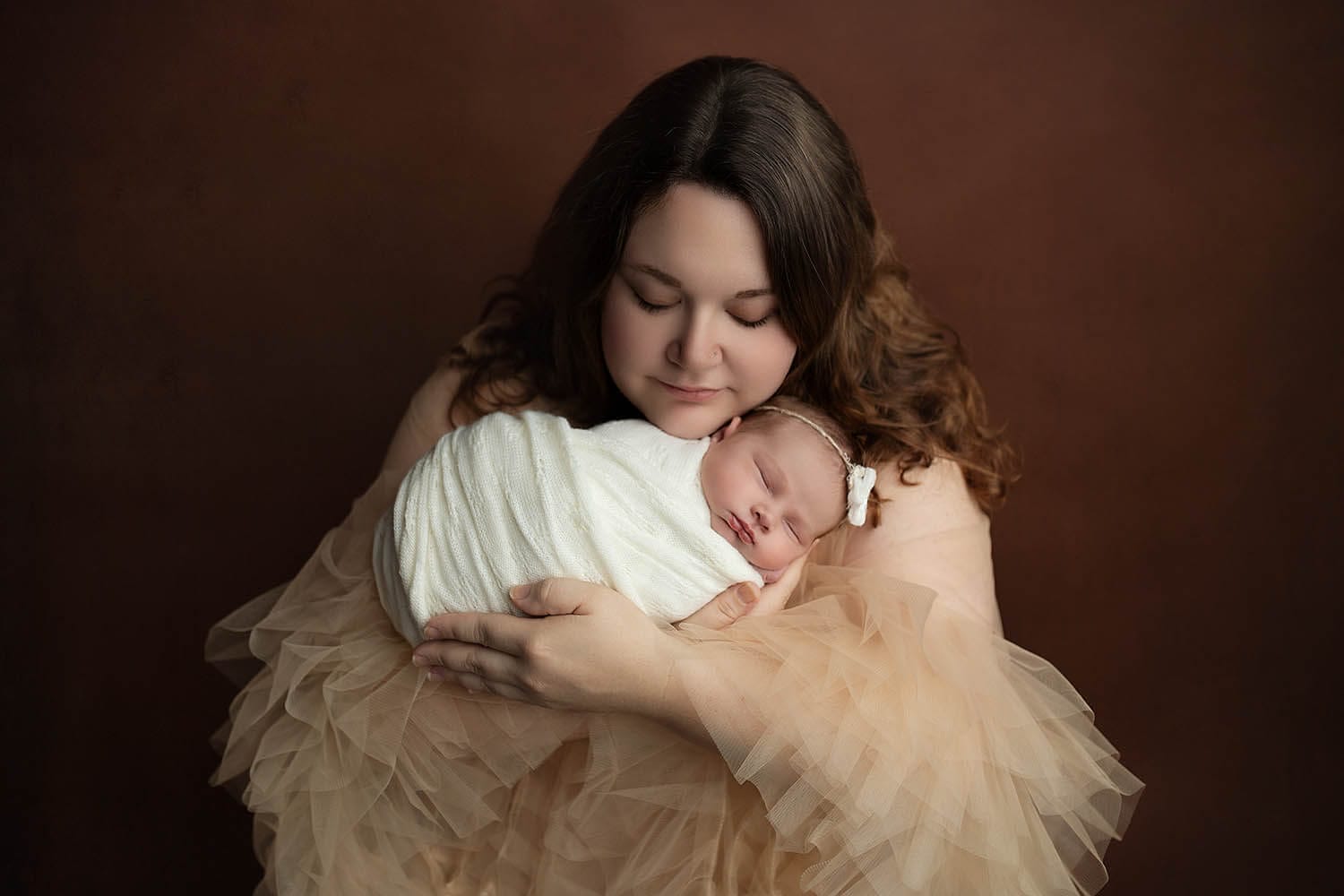 A mother rests her chin on her sleeping newborn baby girl in her hands in a studio