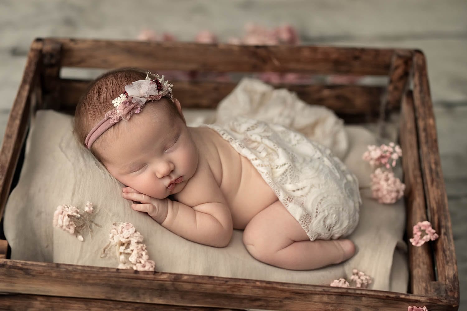A newborn baby sleeps in a wooden crate in a lace blanket and pink headband with flowers around her thanks to lactation consultant new orleans