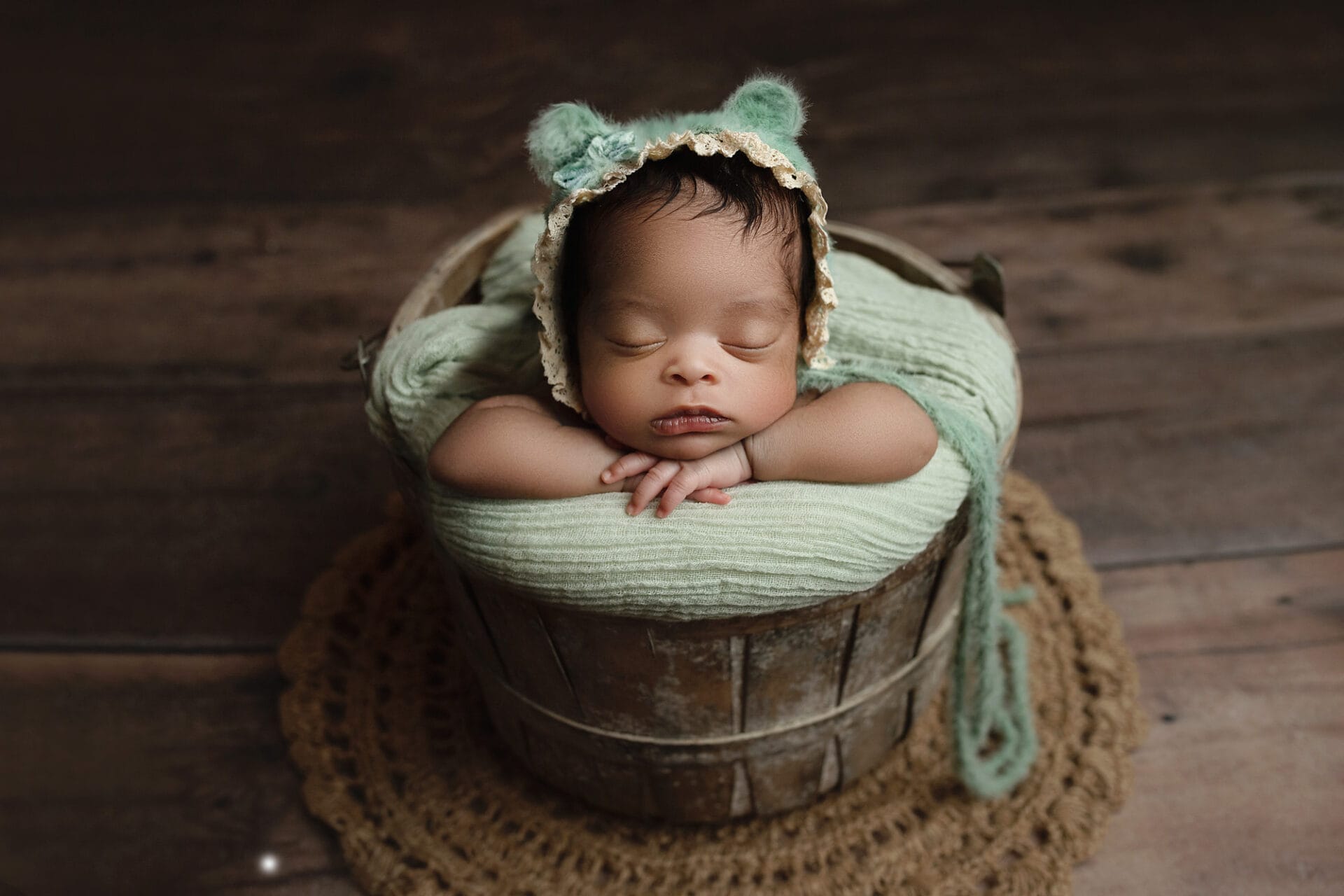 Newborn baby girl with bear bonnet in bucket with hands under chin with New Orleans baby boutique.