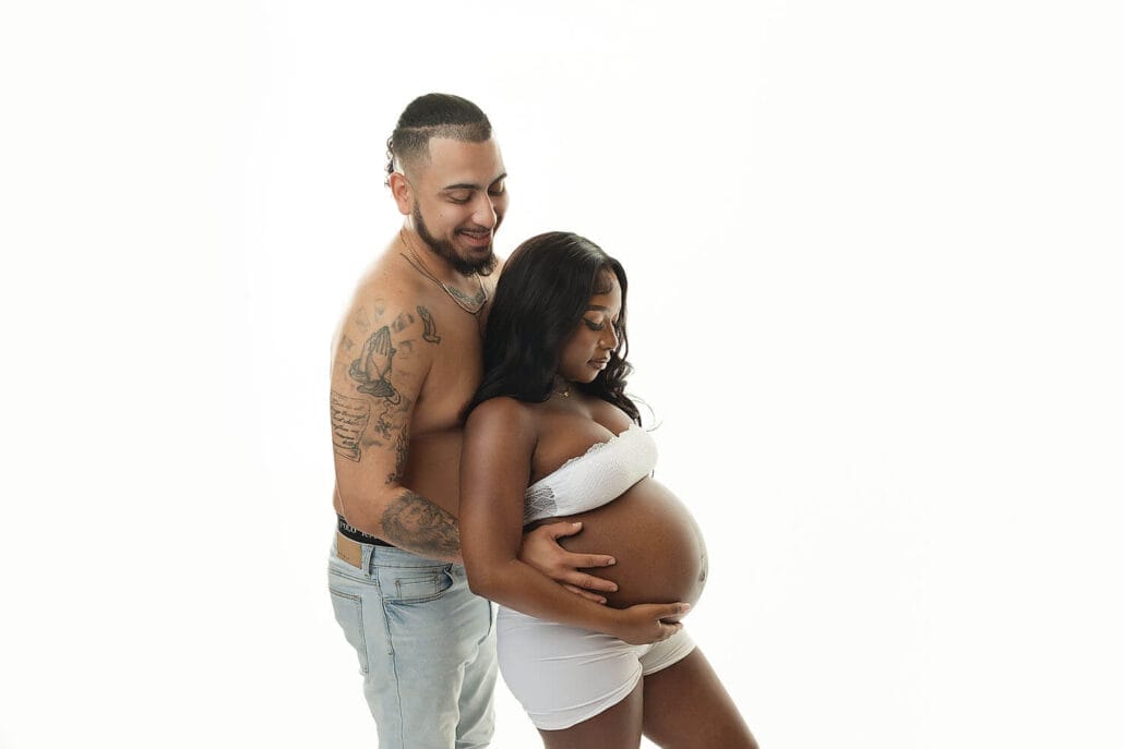 A mother to be in white underwear stands in a studio holding her bump leaning into her shirtless husband