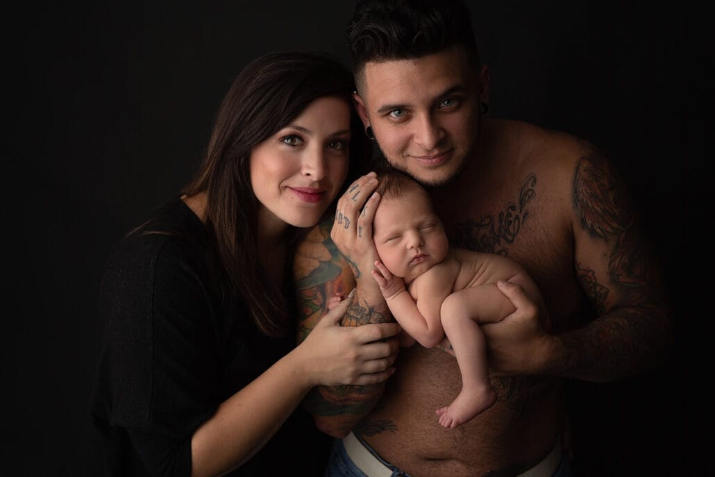 A tattoo covered father cradles his sleeping newborn baby shirtless in a studio with mom hugging onto them after meeting new orleans nannies