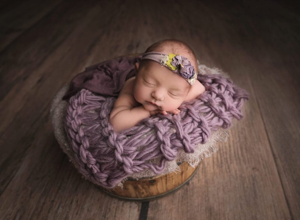 A newborn baby girl in the studio in a wooden bucket with purple layers wearing a flower headband.