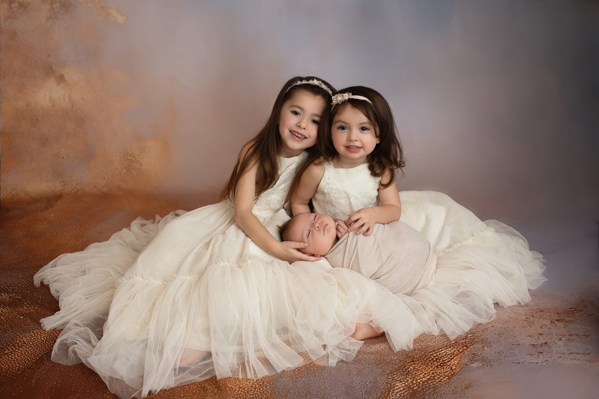Two toddler sisters in matching white dresses sit in a studio with their sleeping newborn sibling in their lap
