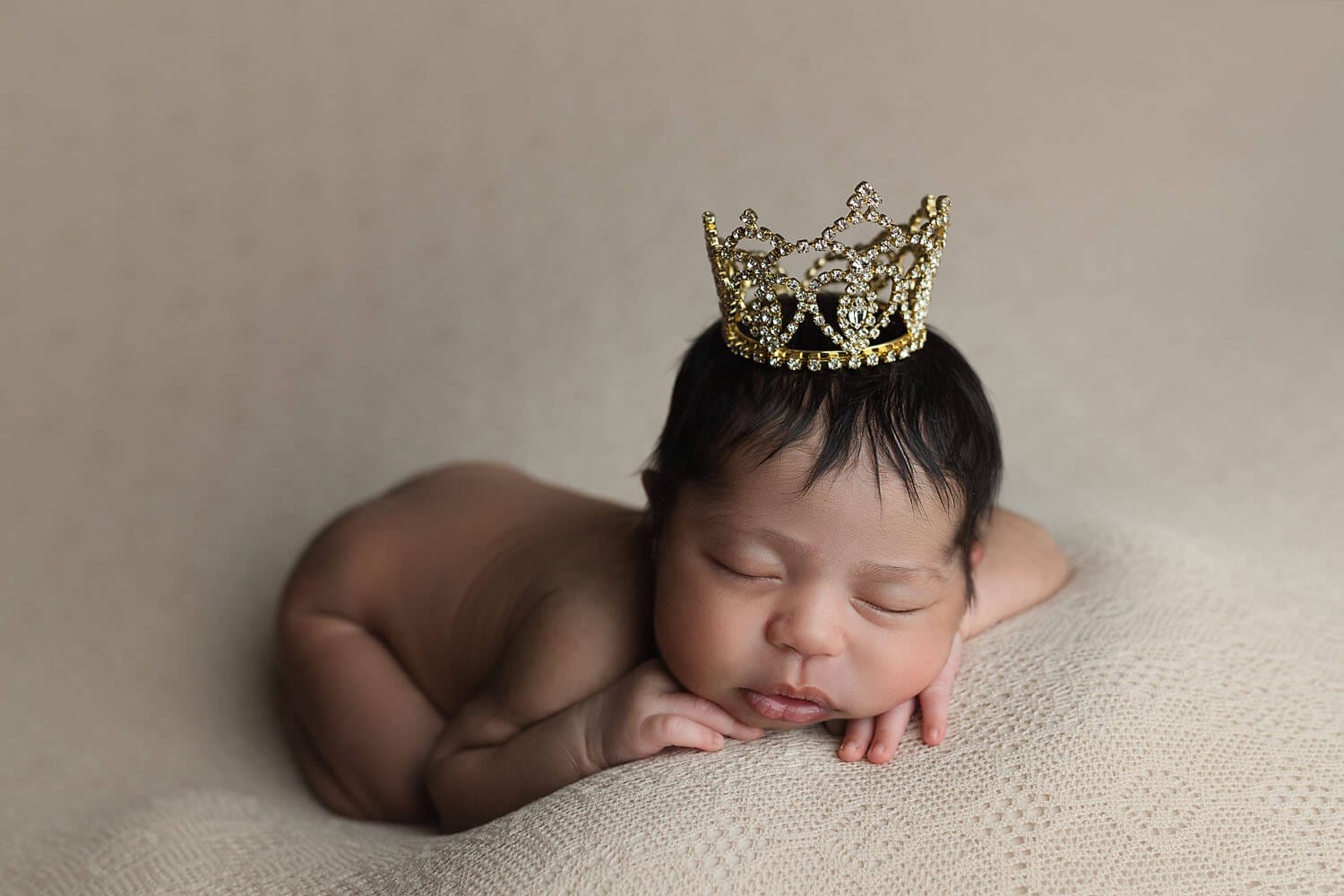 A newborn baby sleeps in froggy pose wearing a gold crown