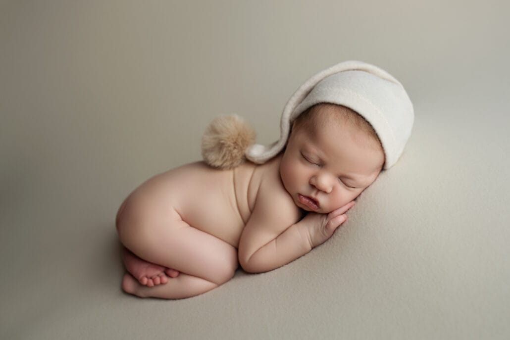 Newborn baby boy in the studio with sleepy hat laying on beige backdrop on the tummy.
