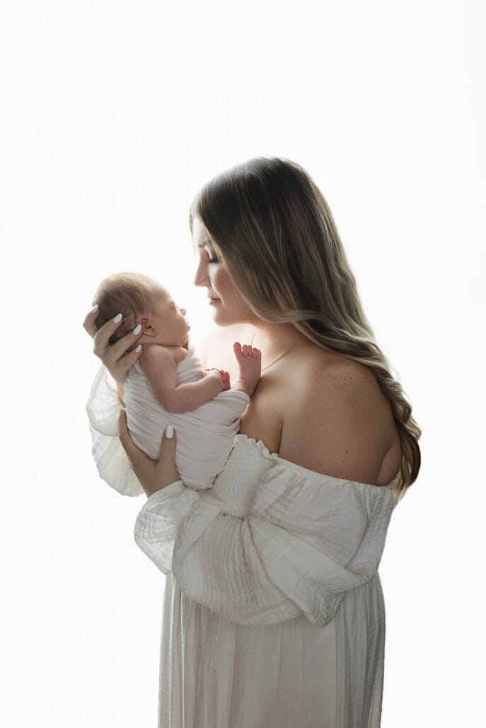 New mom in a white dress with baby boy swaddled in a white wrap in the studio with Baton Rouge lactation consultant.