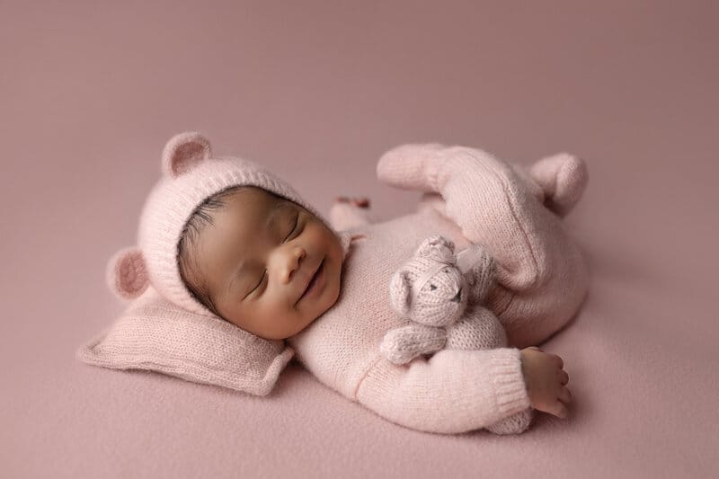 A newborn baby girl in the studio is smiling with a pink bear bonnet and hugging a teddy bear.