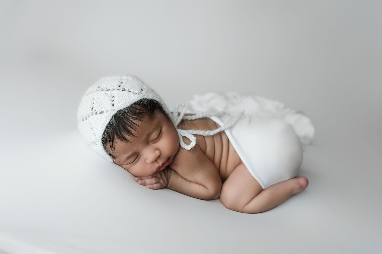 The newborn baby girl is in the studio on the back backdrop, lying on her belly with a white bonnet at the Baton Rouge doula. 