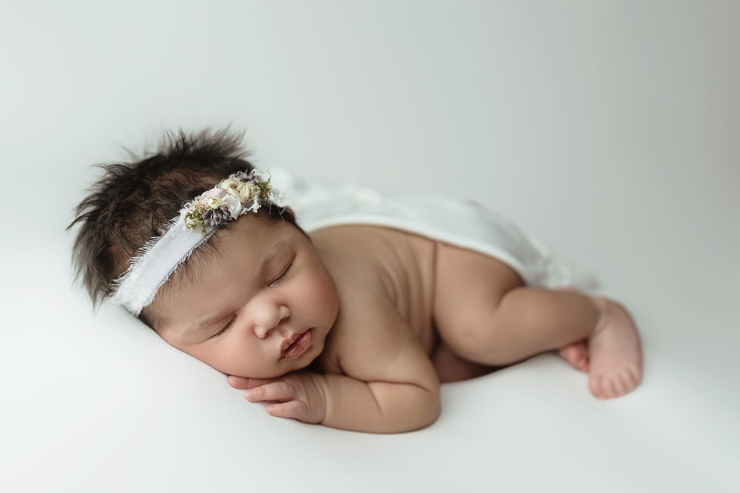Newborn baby girl with white floral headband lying on her tummy with hand under cheek with Baton Rouge doula.