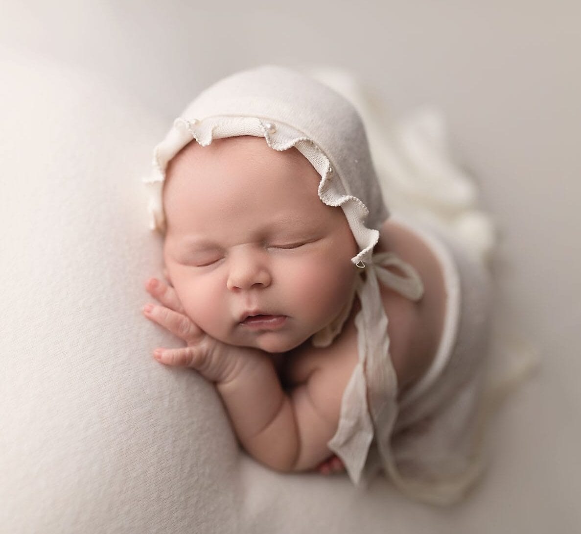 A newborn in the studio with a ivory bonnet with pearls in a taco pose on an ivory backdrop with Baton Rouge prenatal chiropractor.