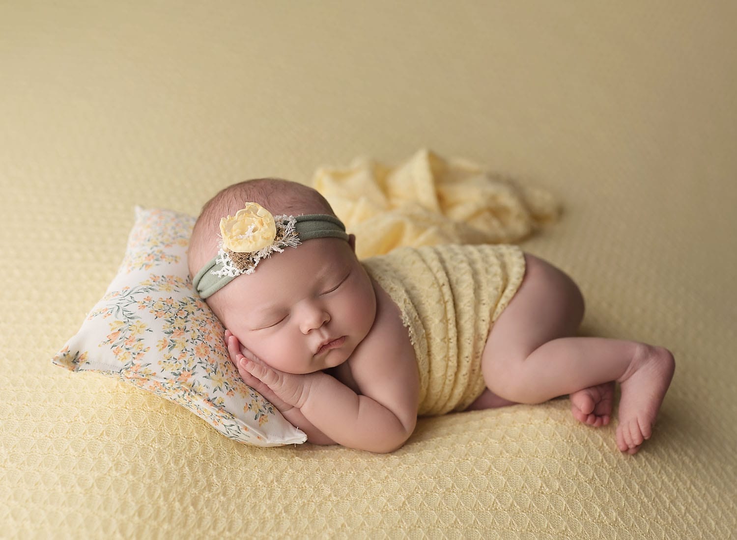 A newborn baby girl in the studio is lying on a yellow backdrop with her head on a floral pillow, wearing a floral headband.
