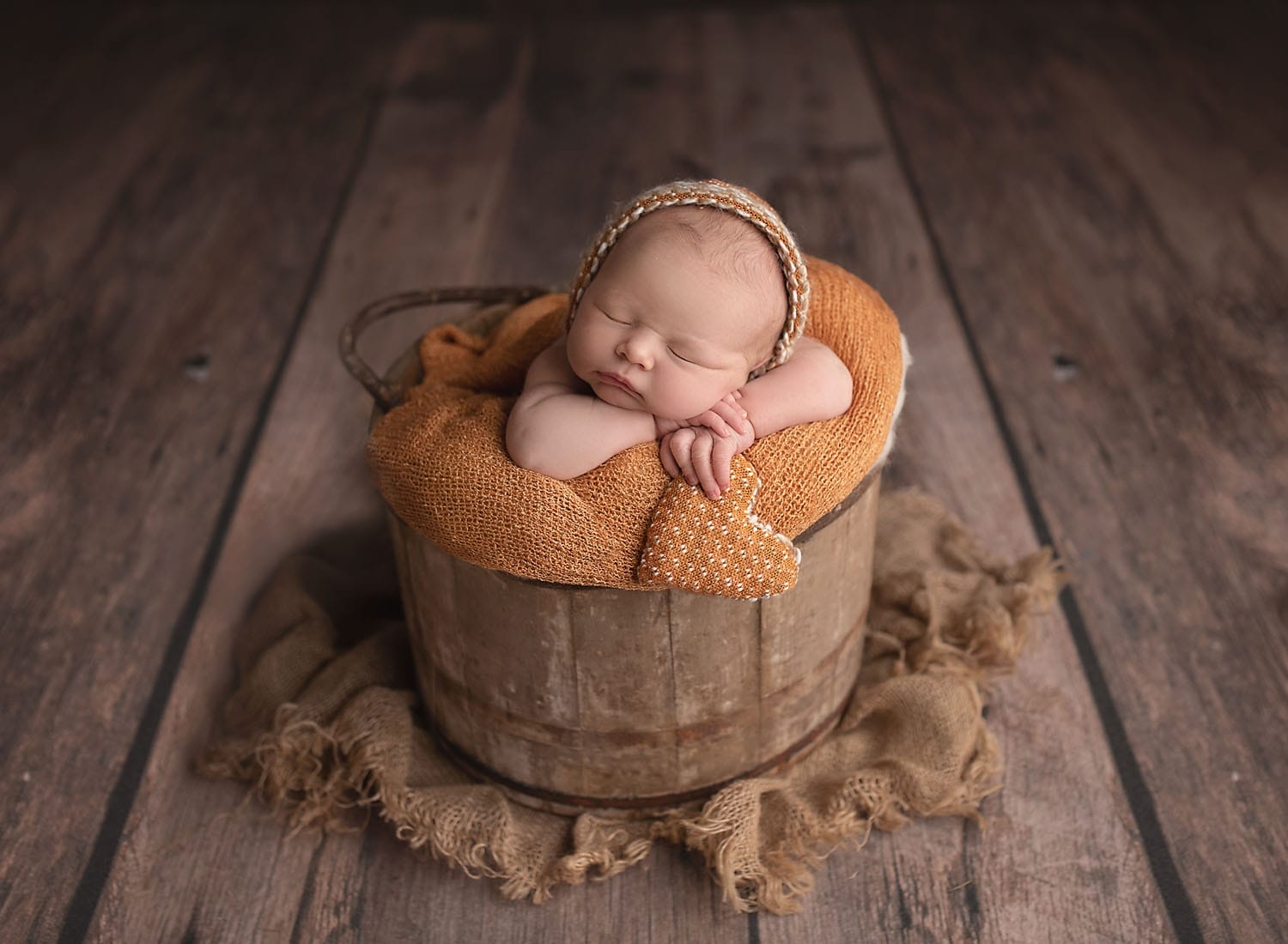 A baby girl in the studio in a wood bucket holding a orange heart pillow at Baton Rouge prenatal chiropractor.