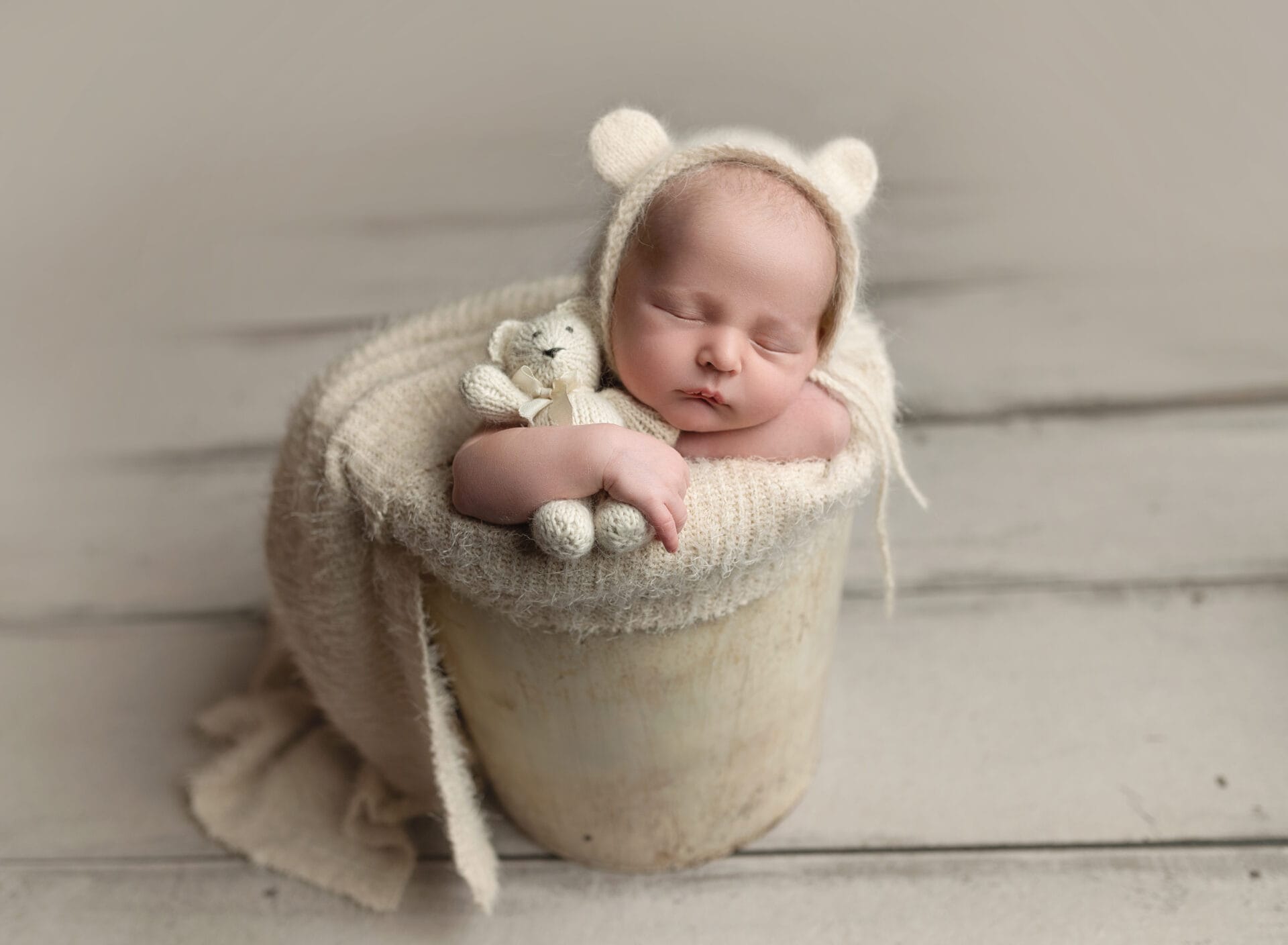A newborn baby in the studio is wearing a bear bonnet in a bucket and cuddling a teddy bear.