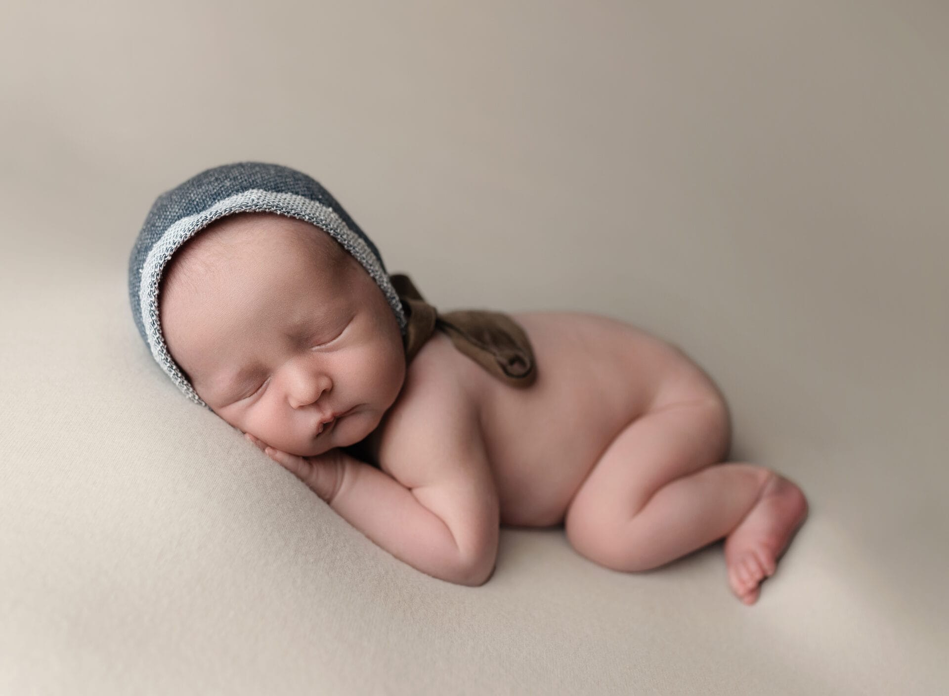 A newborn in the studio with a blue bonnet is lying on a cream backdrop on his belly.