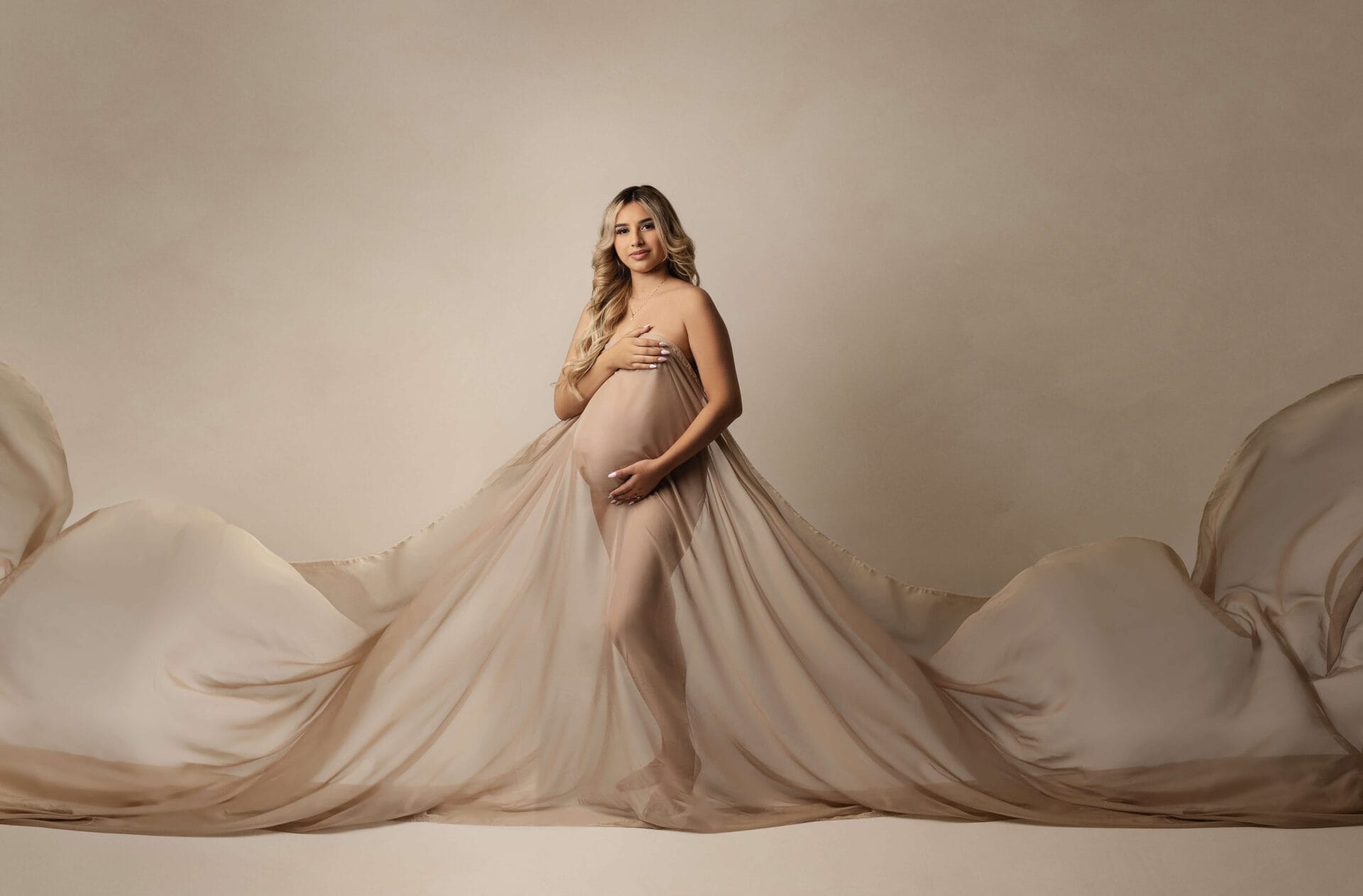 A pregnant mom is to be in the studio with fabric draped, embracing her baby bump. 