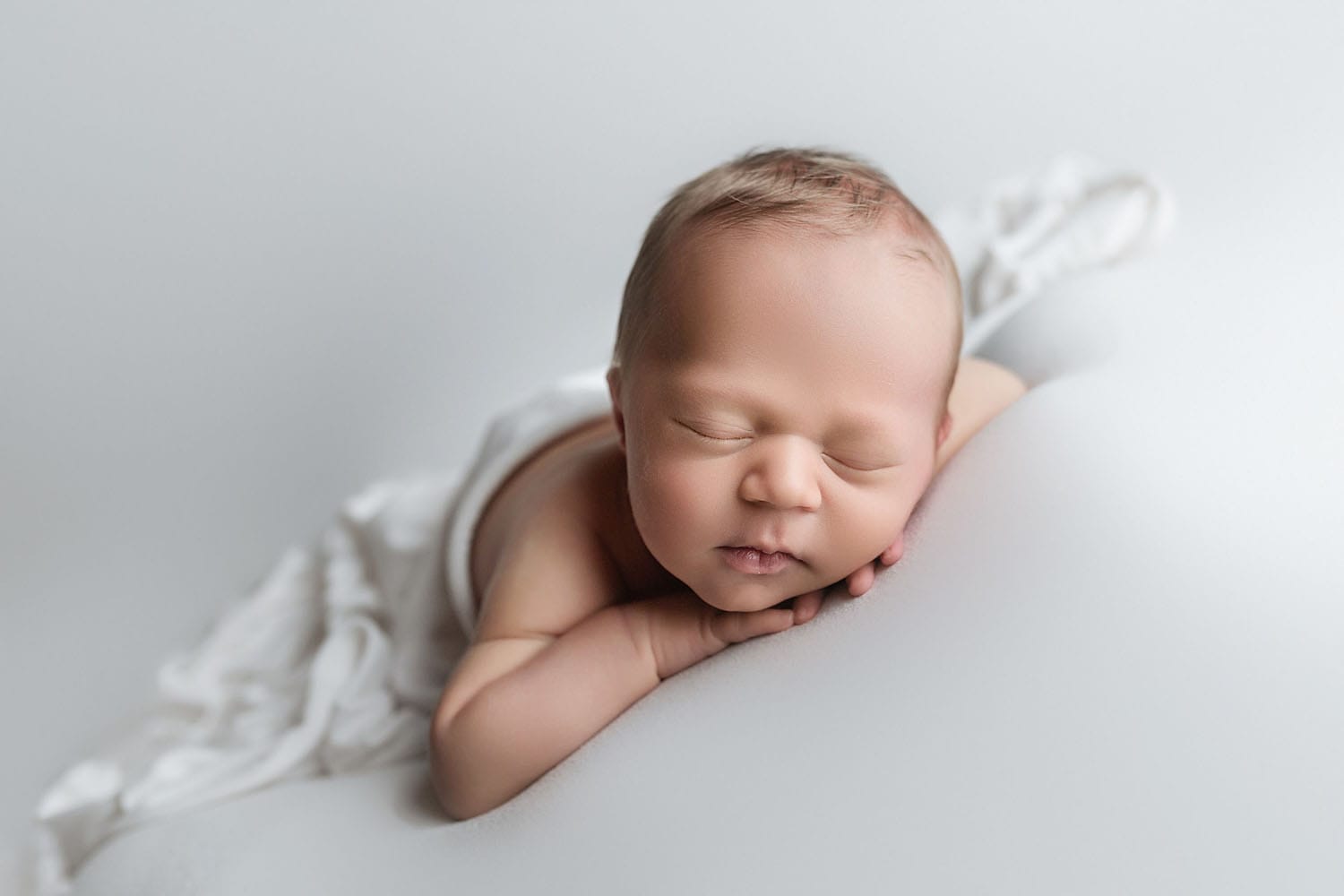 A baby boy in the studio lying on a white backdrop with fabric draped over his back posed with his chin resting on his hands.