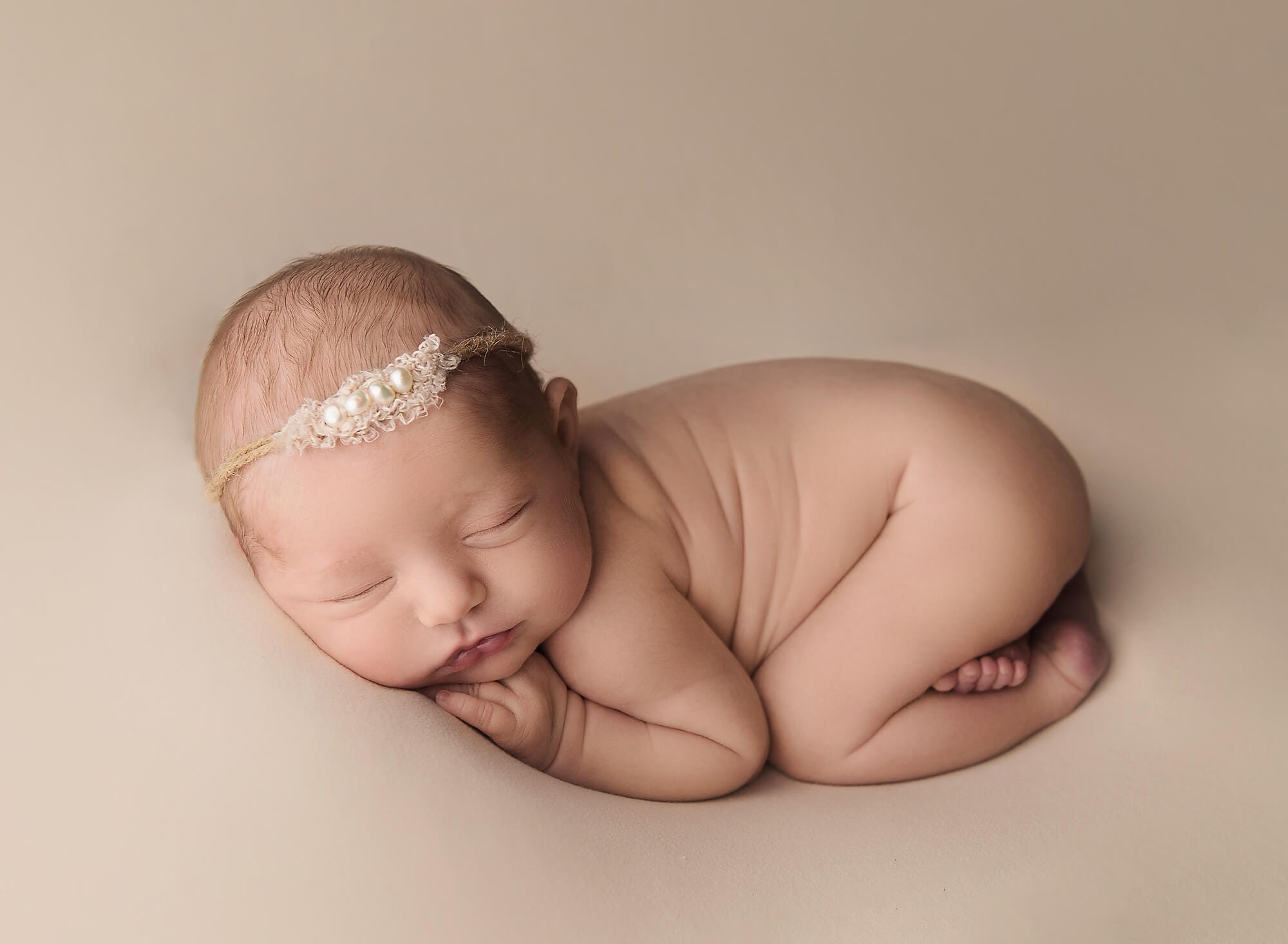 The newborn baby with a dainty headband is lying on a tummy curled up on a beige background in the studio. 