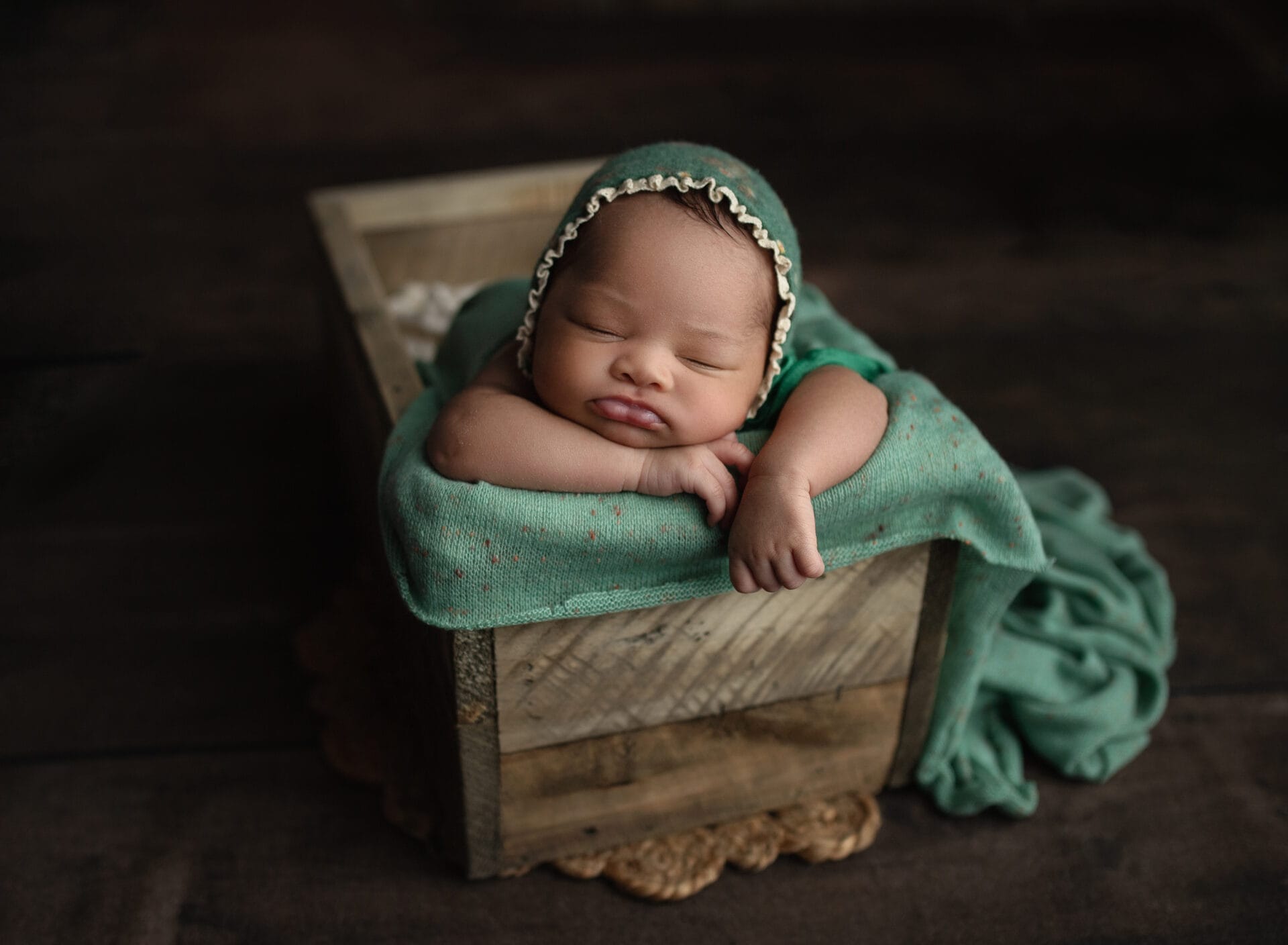 A newborn baby girl in the studio with a mint green bonnet is lying in a crate with her hand under her chin at Lafayette lactation consultant.