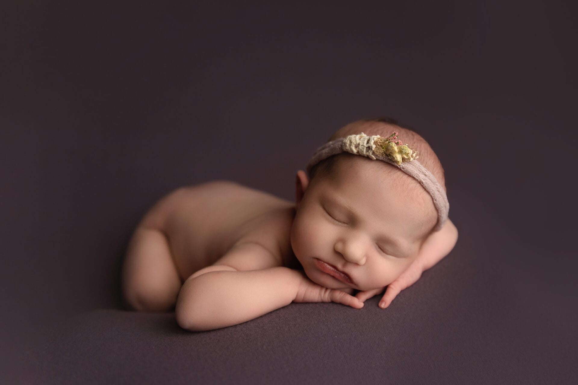 A newborn baby in the studio is wearing a floral headband and lying on a purple backdrop with her head in her hands.