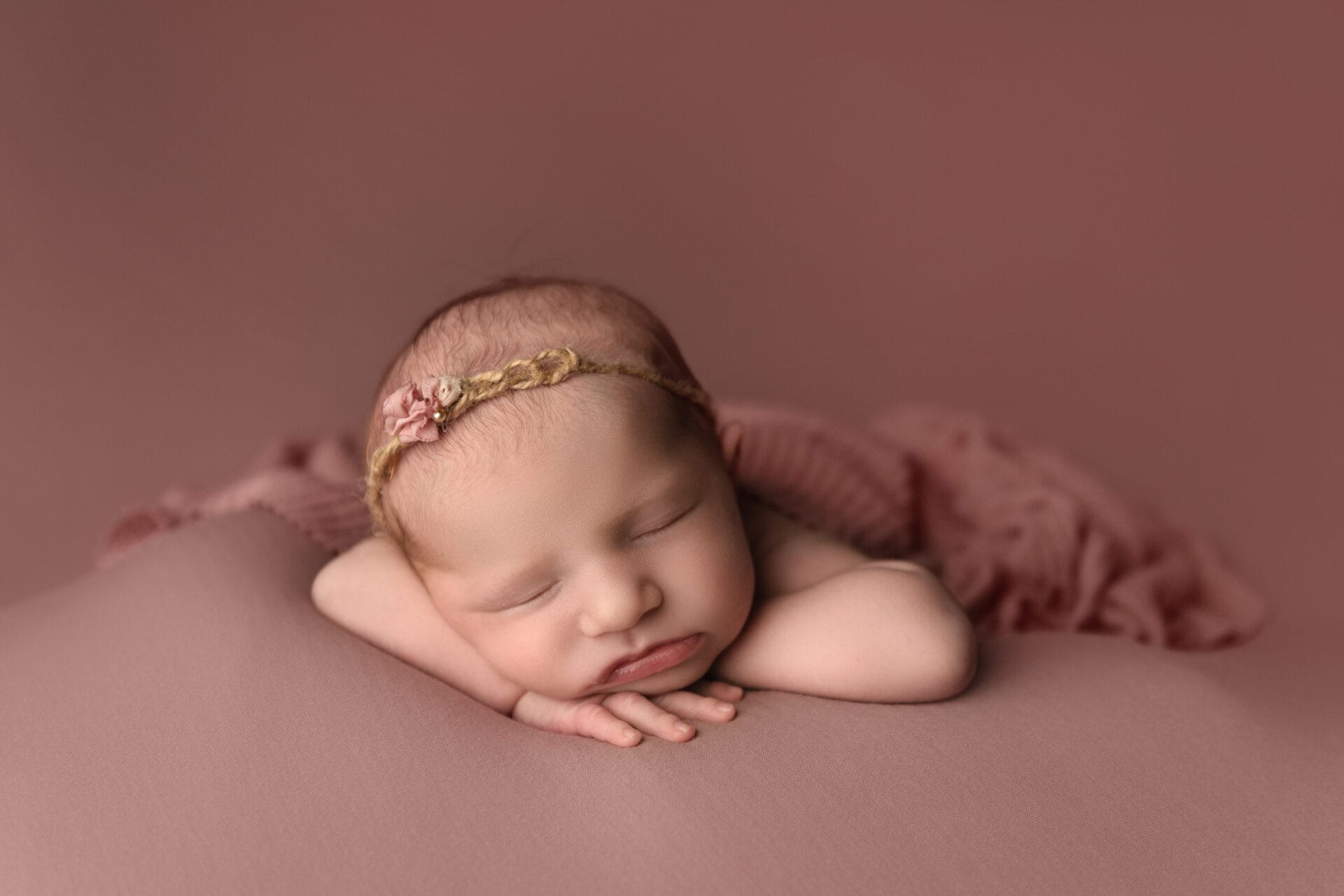 A baby girl in the studio lies on a pink backdrop, a wrap draped over her back, resting her head on her hands, wearing a pink flower headband.