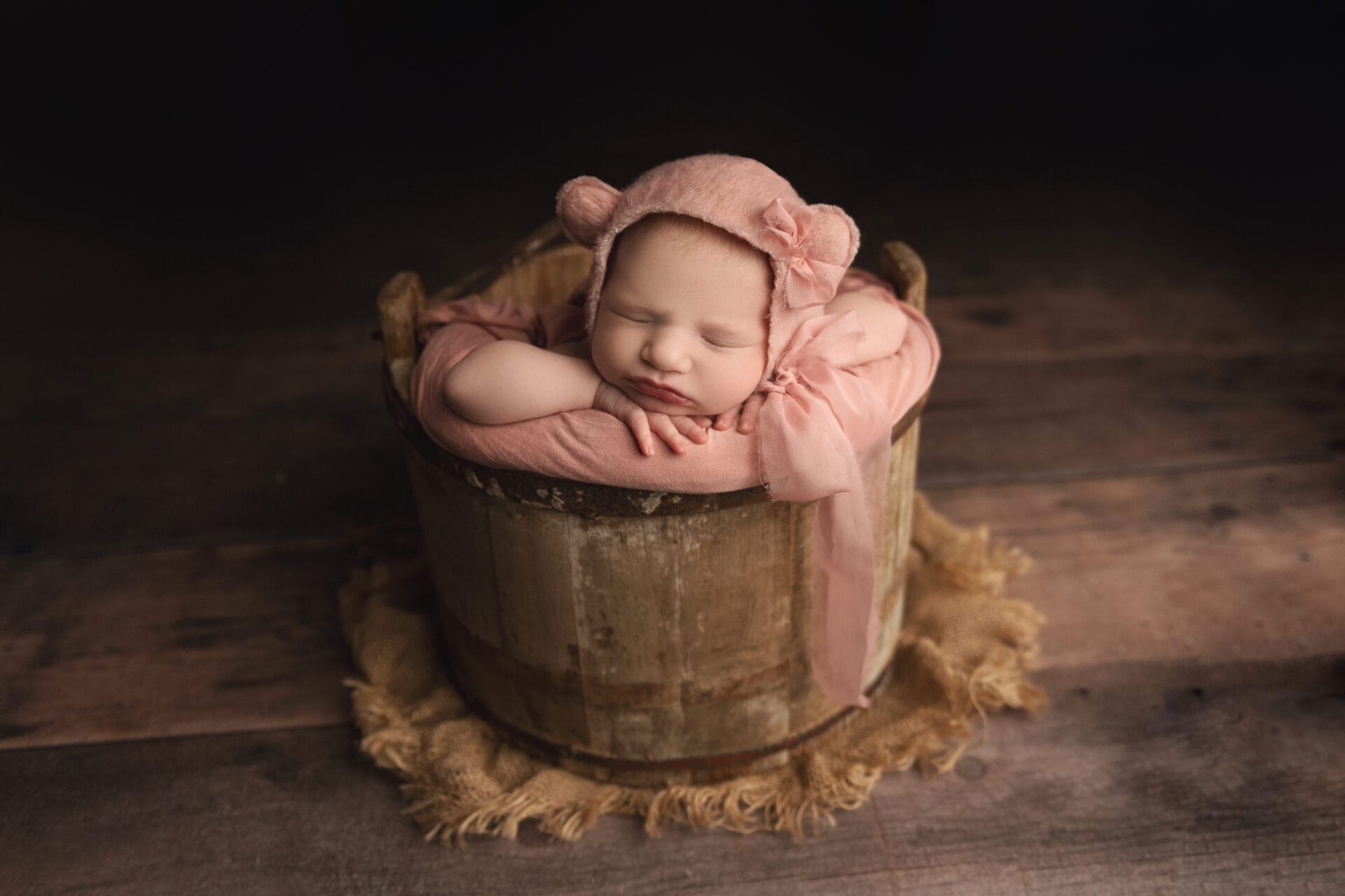 A baby girl in a wood bucket is wearing a pink bear bonnet, resting her chin on her hands with Lafayette's top daycare.