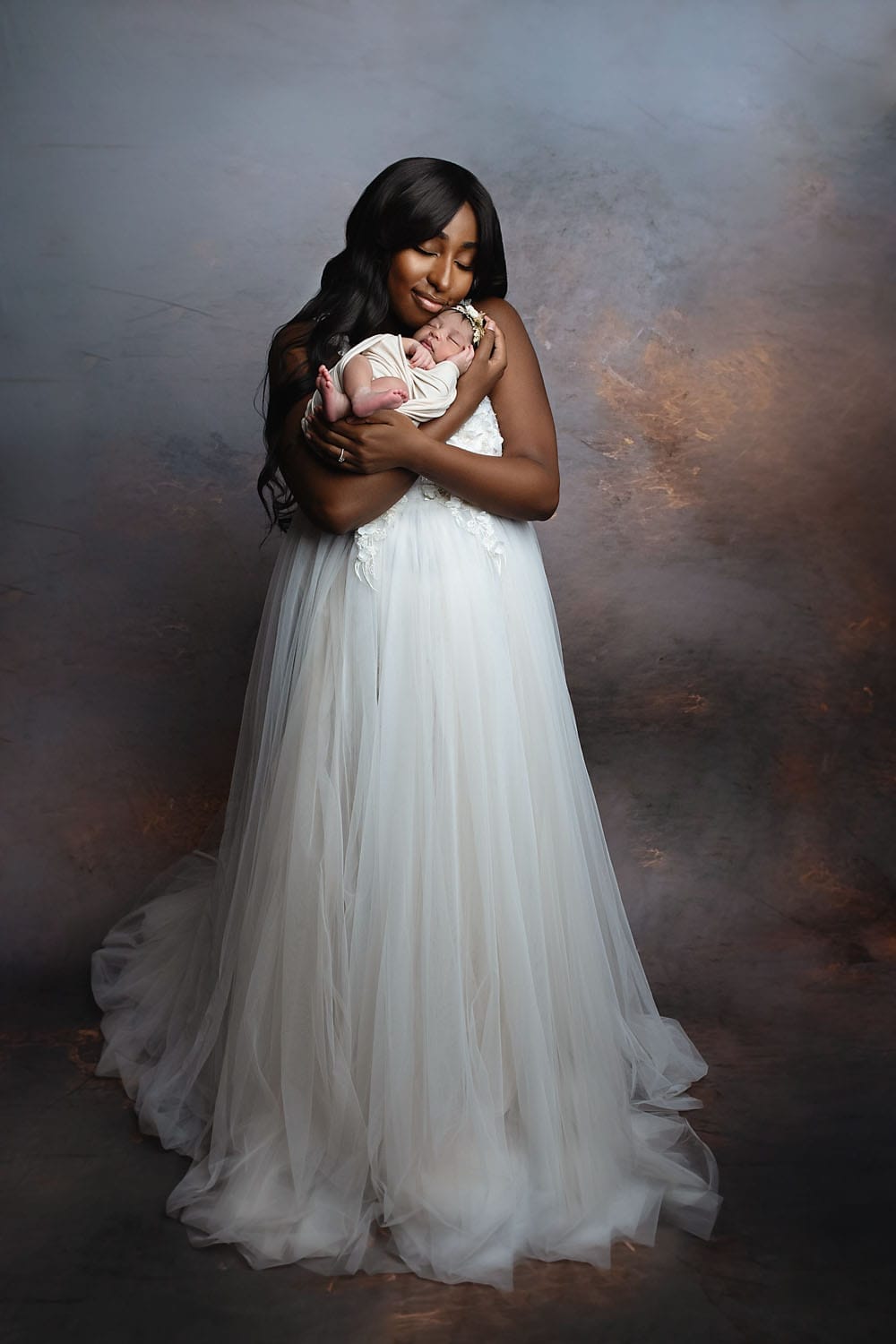 A new mom is holding her baby girl in the studio in an elegant white gown at New Orleans OBGYN.