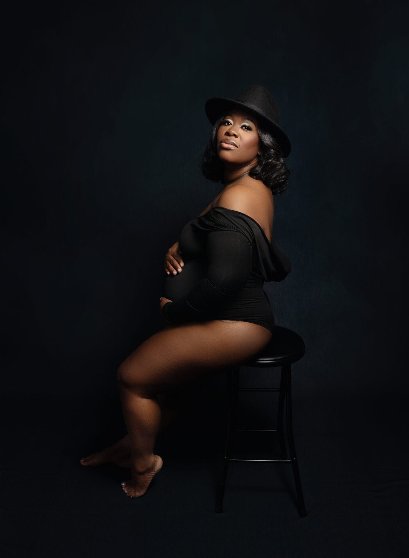 An expectant mom in the studio is sitting on a stool with a black body suit and black hat, embracing her pregnant belly at New Orleans prenatal yoga studio. 