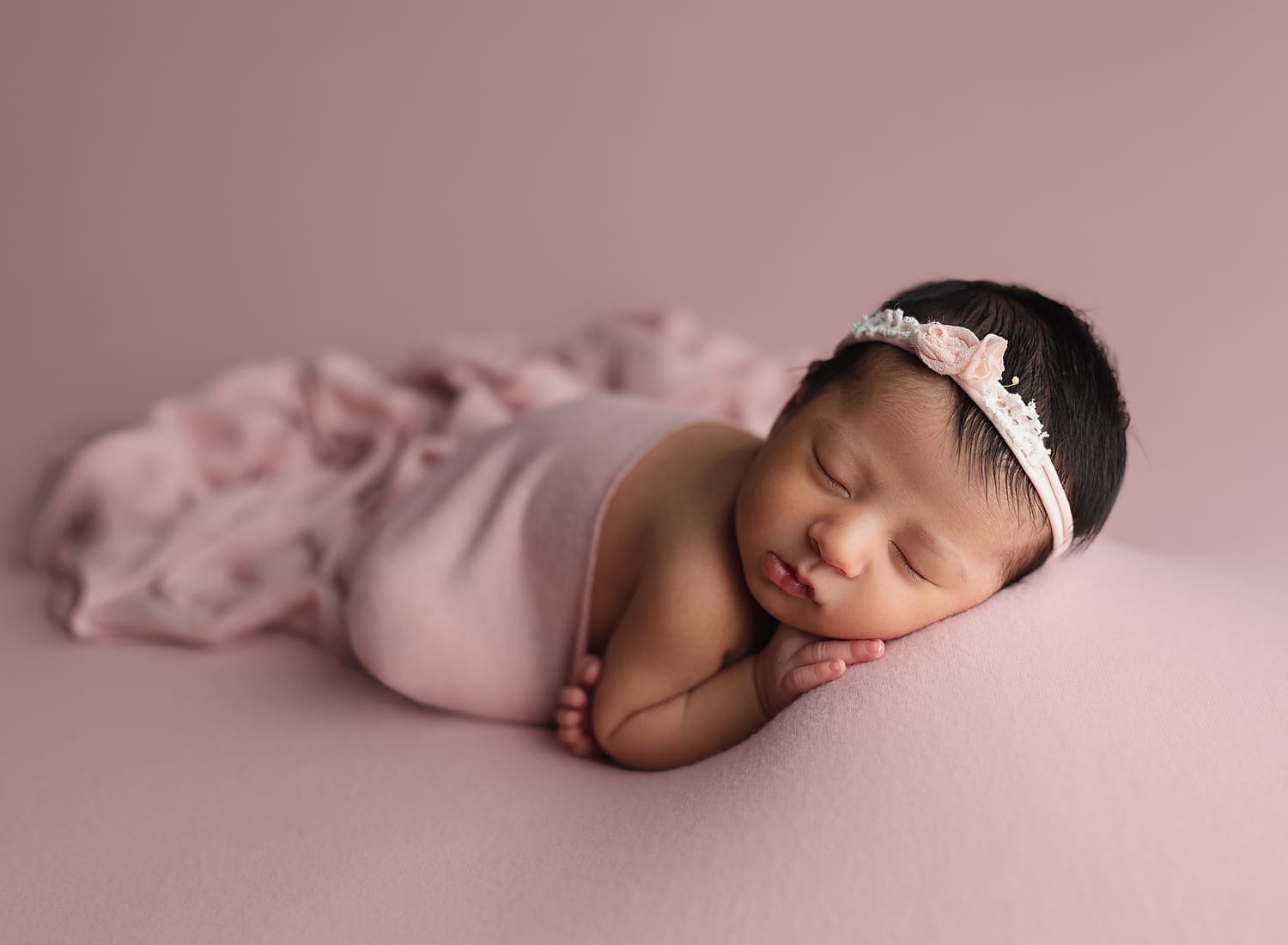A newborn baby girl in the studio lying on a pick backdrop with a pink floral headband with feet curled up.