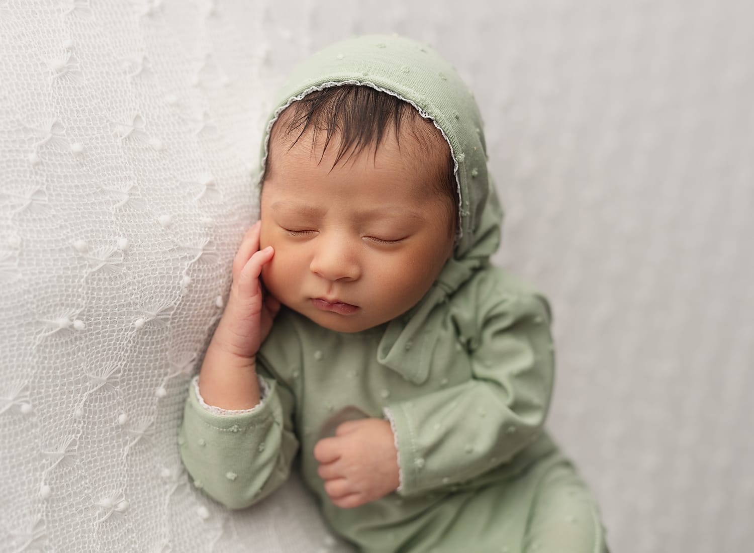 A baby girl in the studio with a green onesie and bonnet laying on a white backdrop at New Orleans's top daycare.