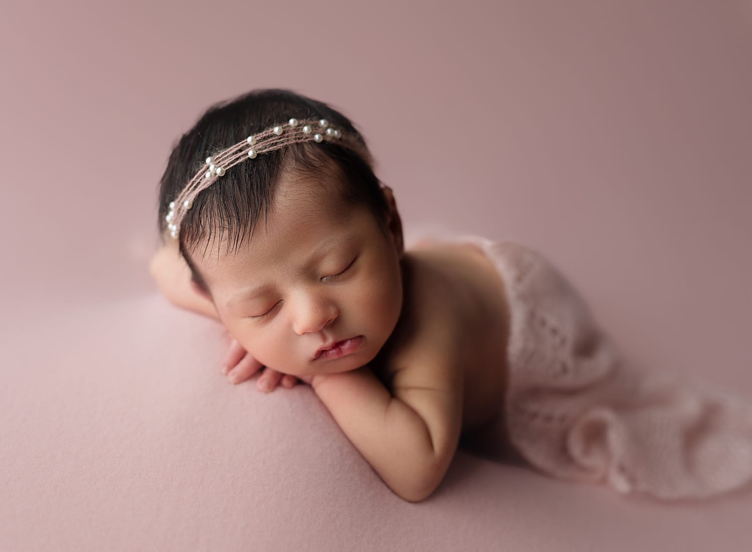 A baby girl in the studio lying on a pink backdrop with a pink lace wrap draped her back with a pearl headband at New Orleans top daycare.