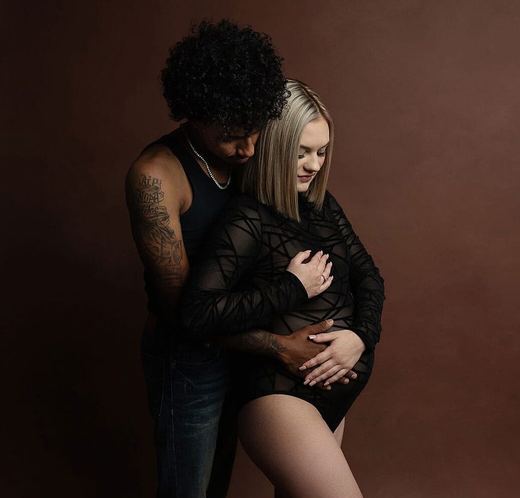A new mom and dad to be in the studio with pregnant mom wearing a black body suit and dad to be is embracing mom's pregnant body.