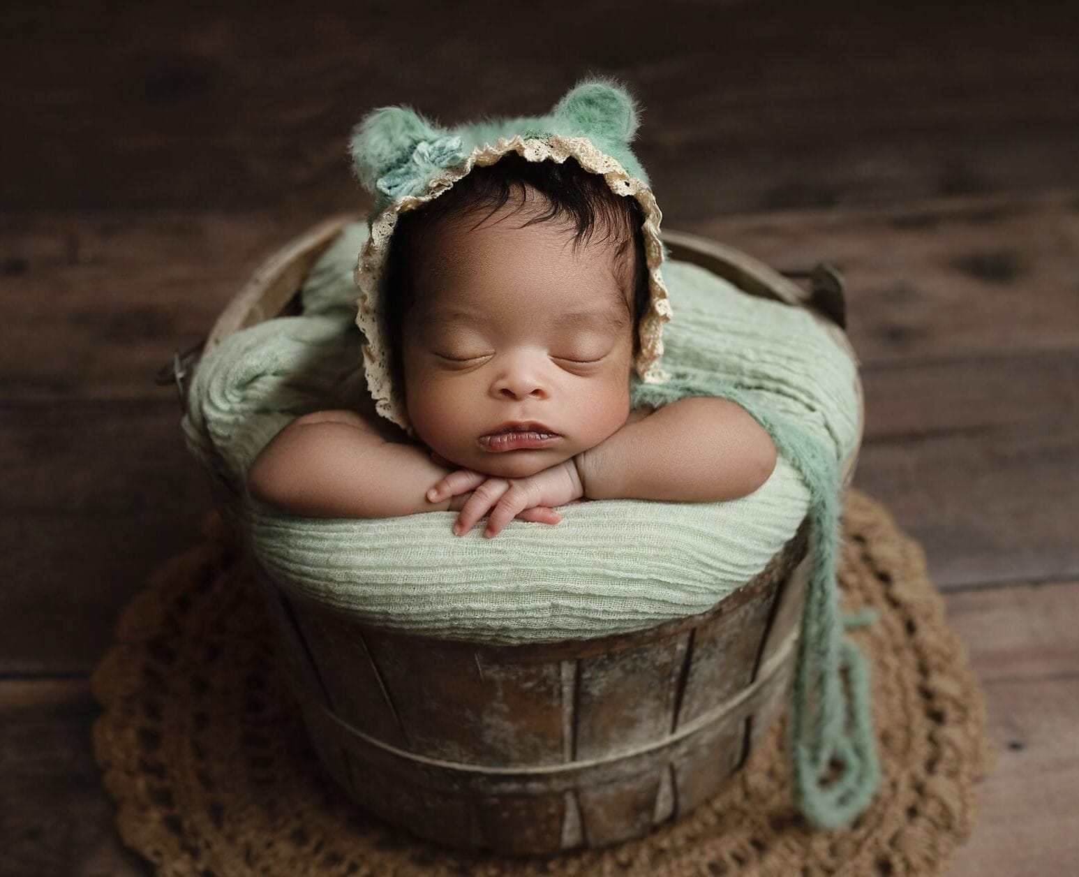 A newborn baby girl in the studio wearing a colored bear bonnet, sitting in a rustic bucket, with hands under her chin at a Lafayette baby shower venue.