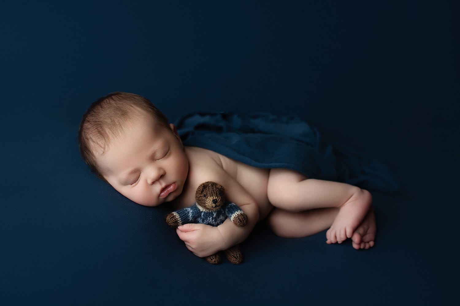 A newborn baby in the studio on a navy blue backdrop, lying on his side, cuddling with a bear. 