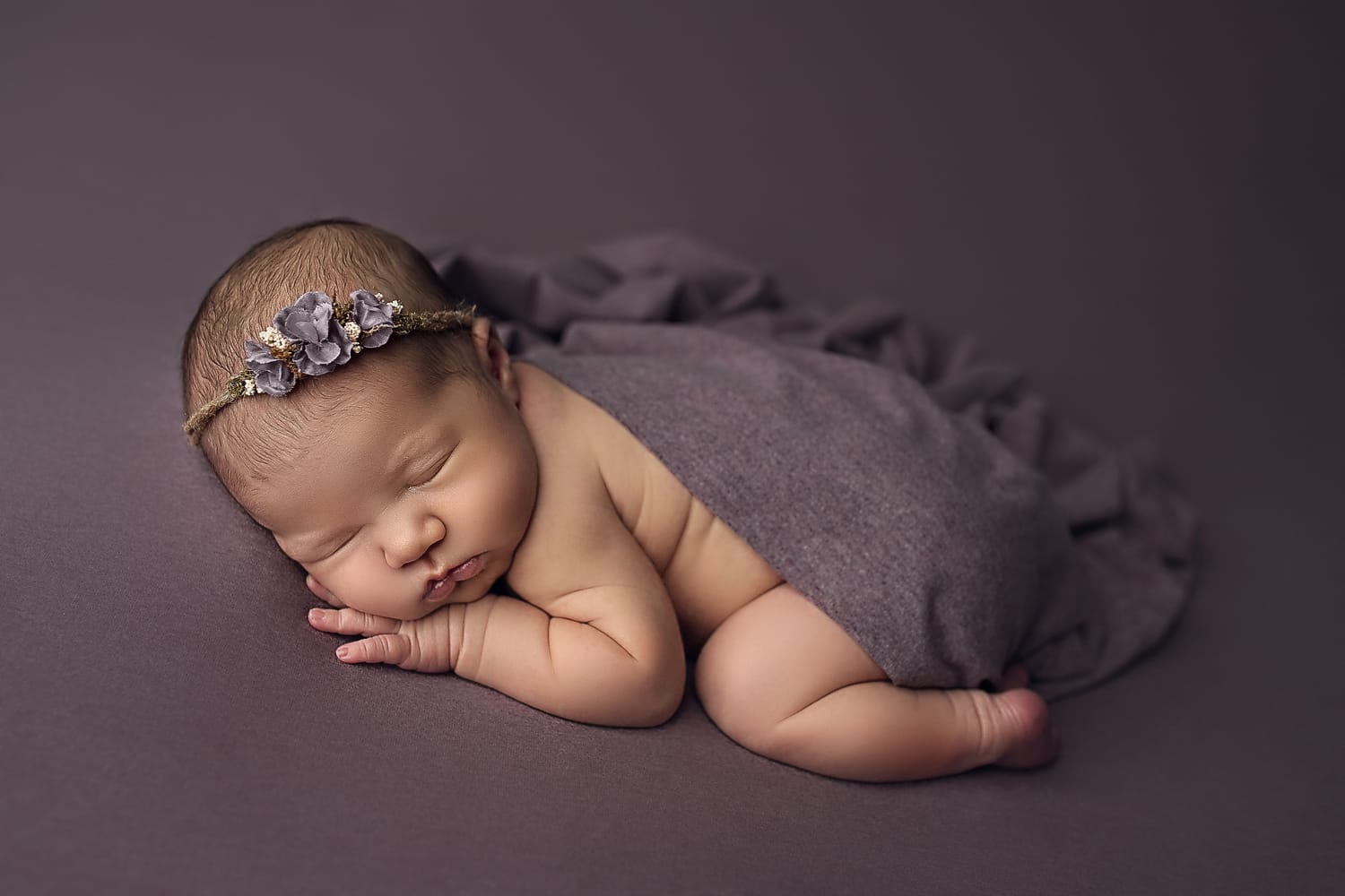 A newborn baby girl is on her tummy on a purple backdrop wearing a floral headband, Baton R, in the studio of auge nanny service. 