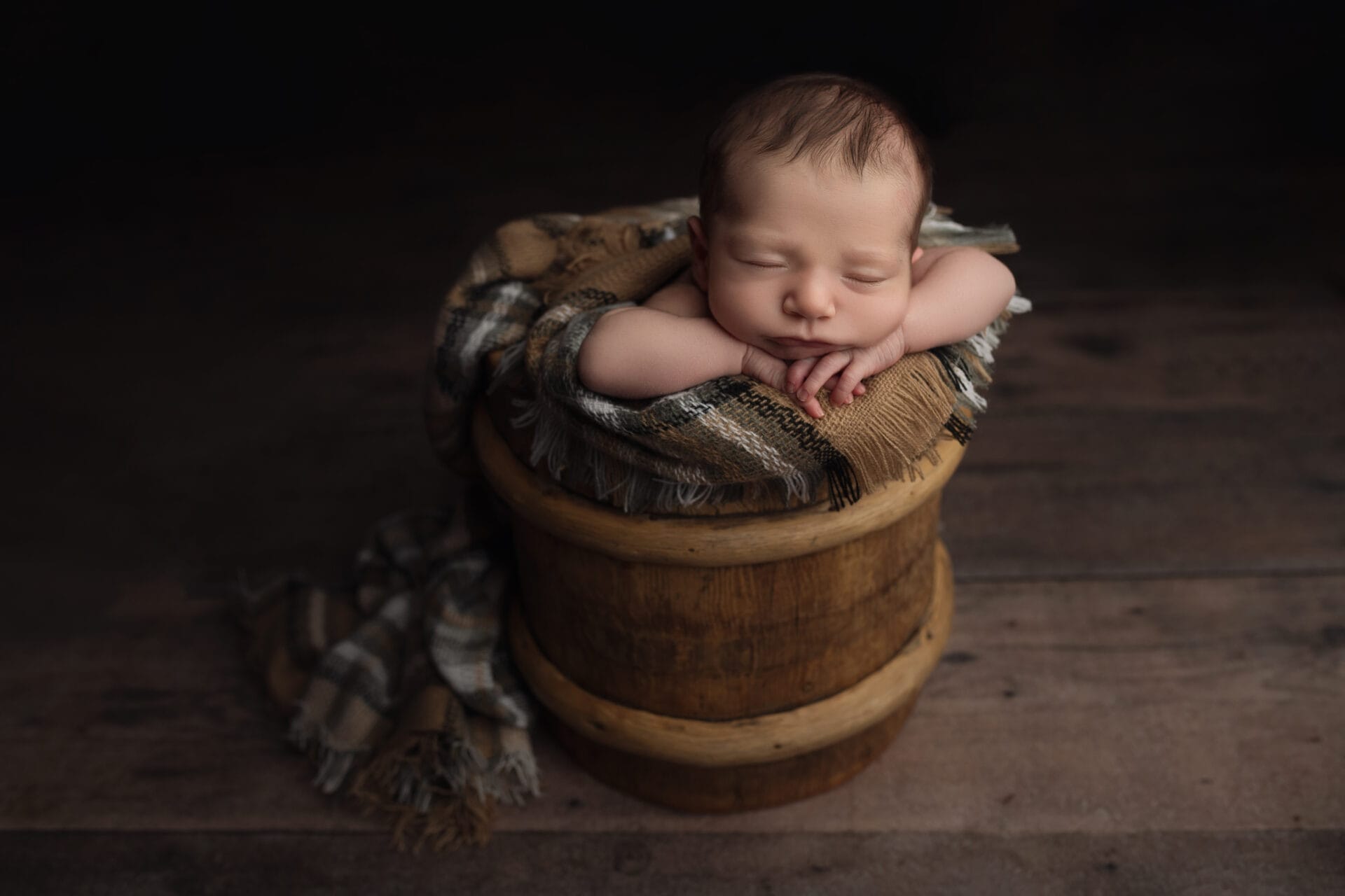A baby boy in the studio with his head on his hands in a wooden bucket with a plaid layer. 