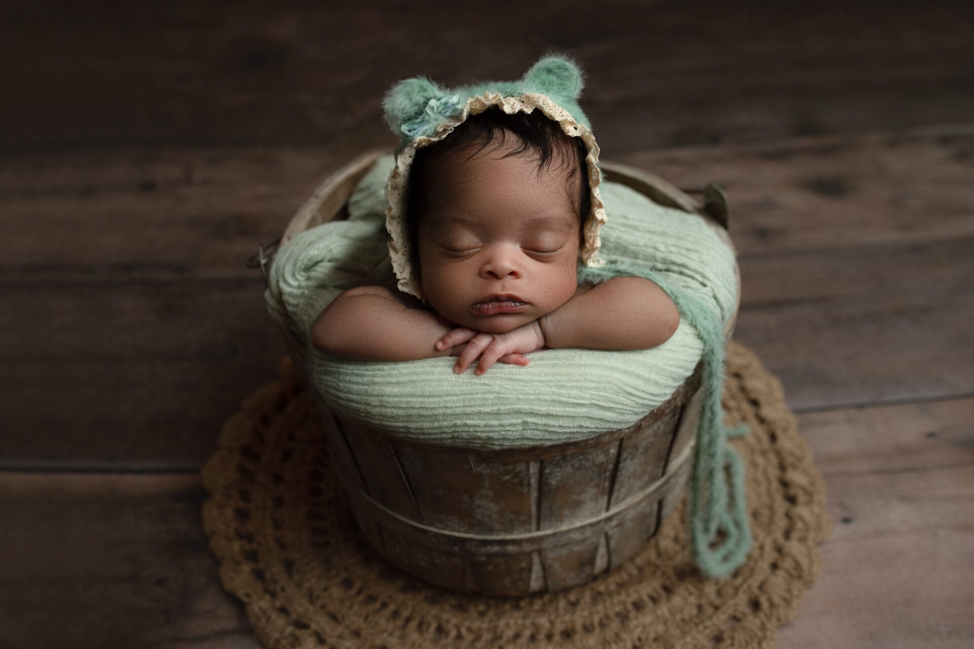 A newborn baby girl in a wooden bucket wearing a mint green bear bonnet. 