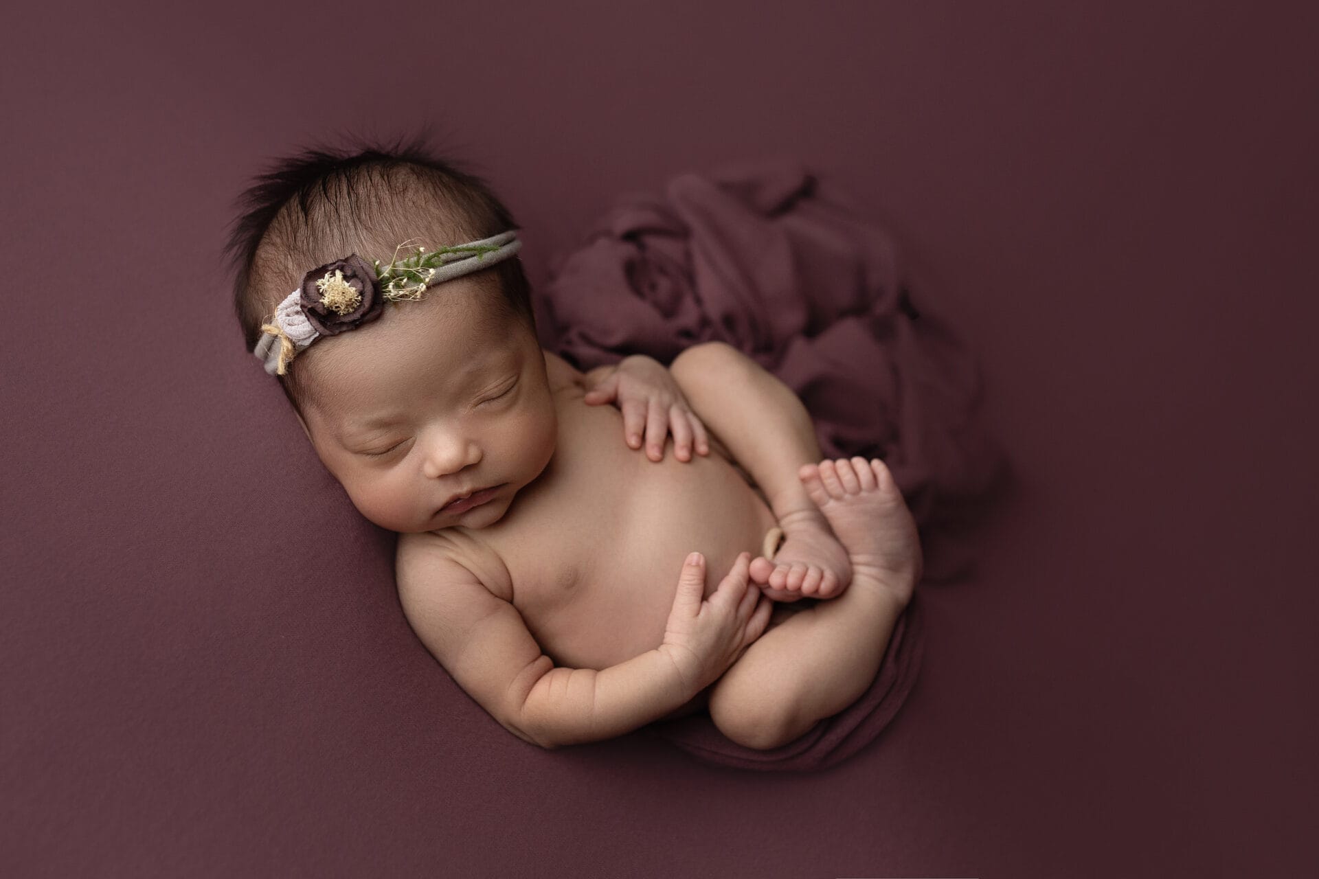 A newborn baby girl in the studio curled up in a womb pose with a floral headband. 